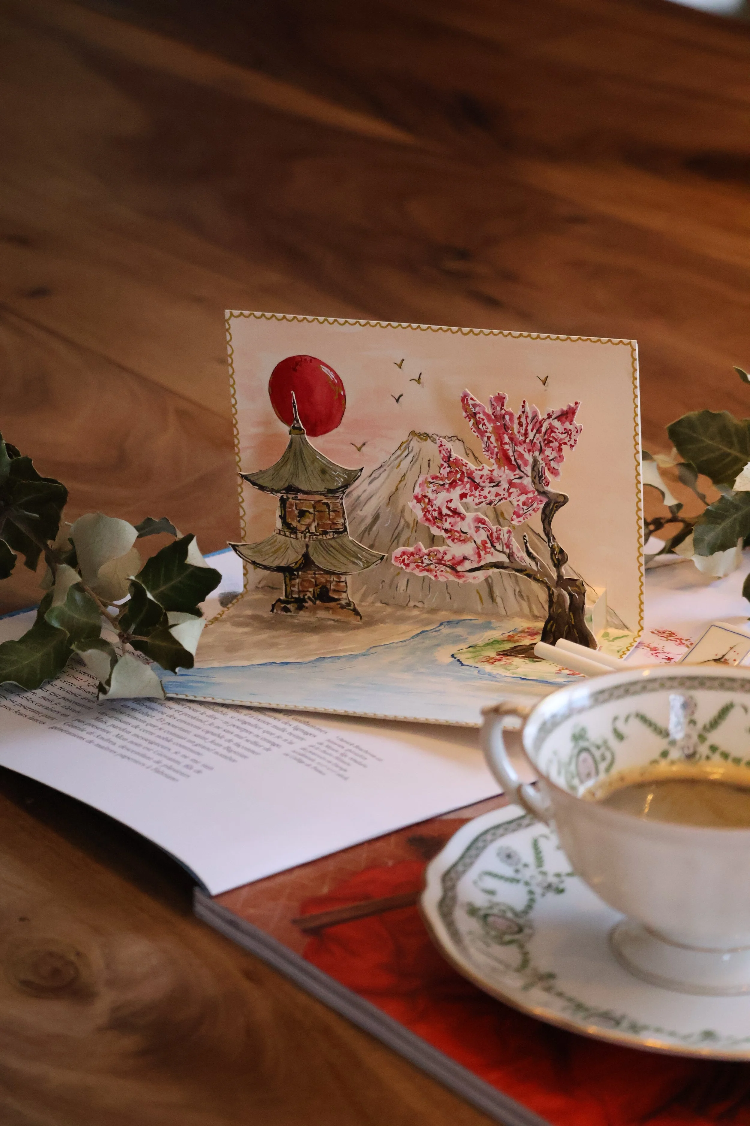 Hand-painted watercolor greeting card with an illustration of a Japanese landscape including a pagoda, cherry blossom tree, mountain, and red sun, placed on a wooden table with an open book, a sprig of holly, and a teacup filled with tea.