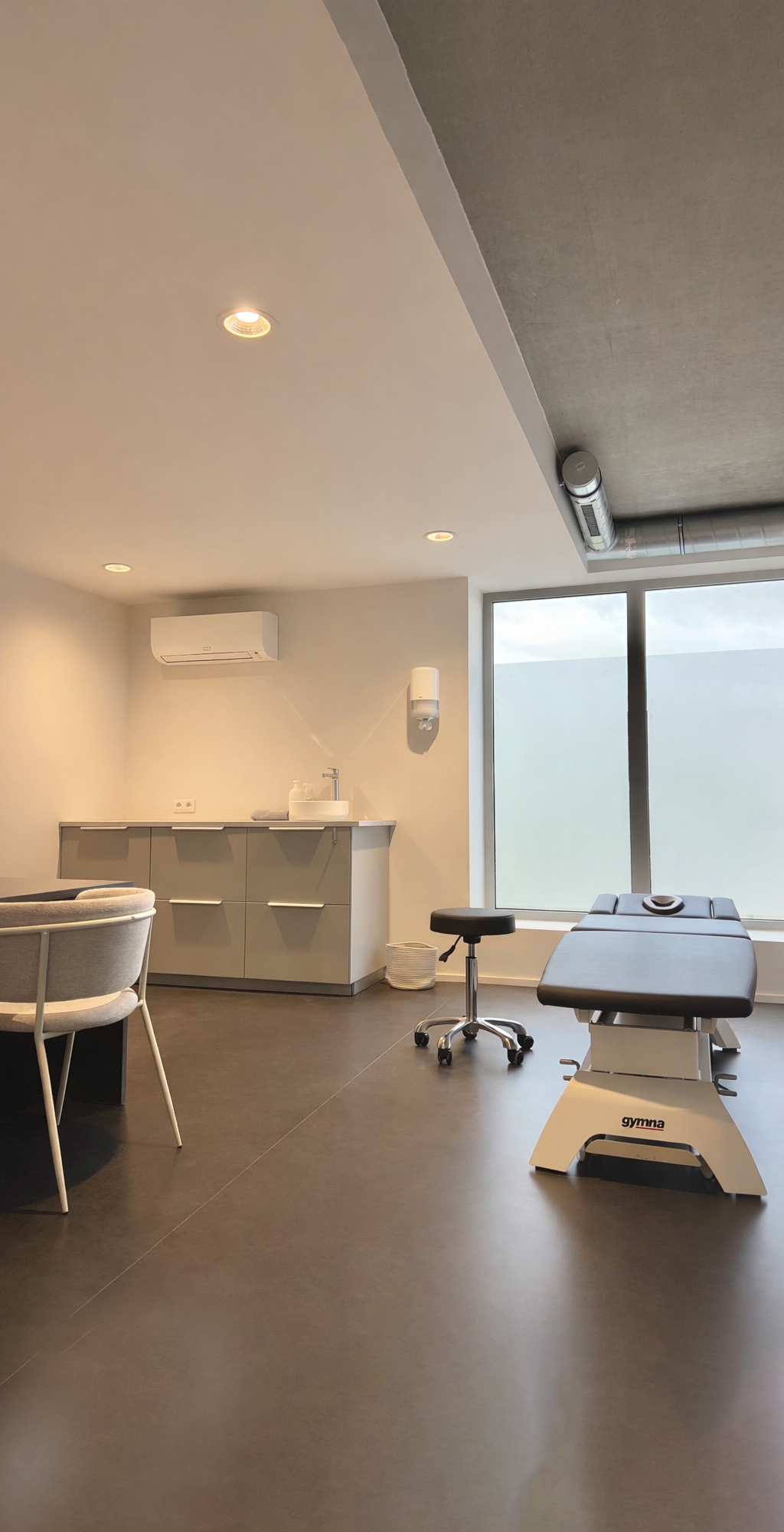 Salle de physiothérapie avec un lit d'examen, un fauteuil, un lavabo, un distributeur de savon, des fenêtres, et un environnement moderne et minimaliste.