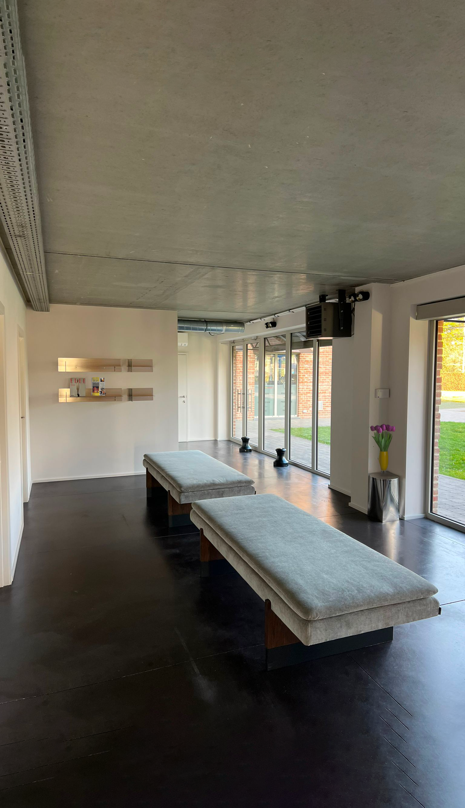 Salon lumineux avec deux bancs recouverts de tissu gris clair, grande baie vitrée, plantes, étagères murales et moquette au plafond.