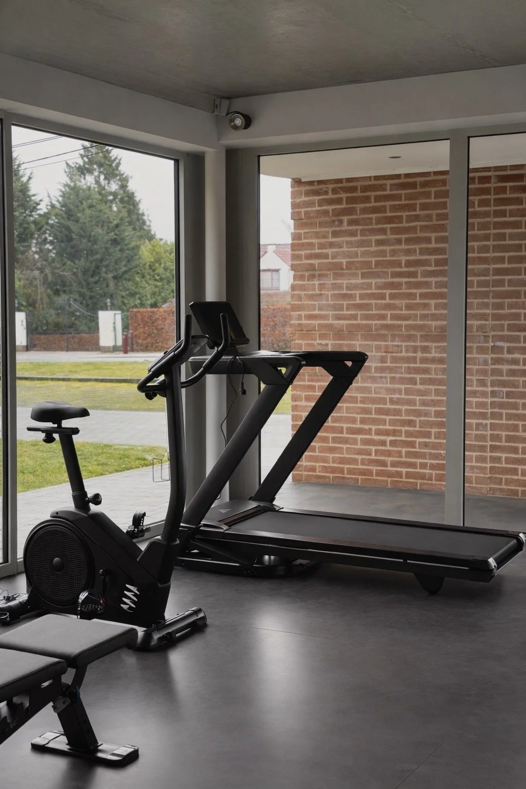 Salle de sport intérieure avec tapis de course, vélo d'appartement et grande fenêtre laissant entrer la lumière naturelle.