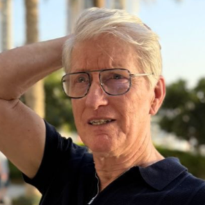 Homme âgé avec des cheveux gris clair, portant des lunettes et un t-shirt noir, souriant, avec un bras derrière la tête, en extérieur avec des arbres et un ciel bleu en arrière-plan.