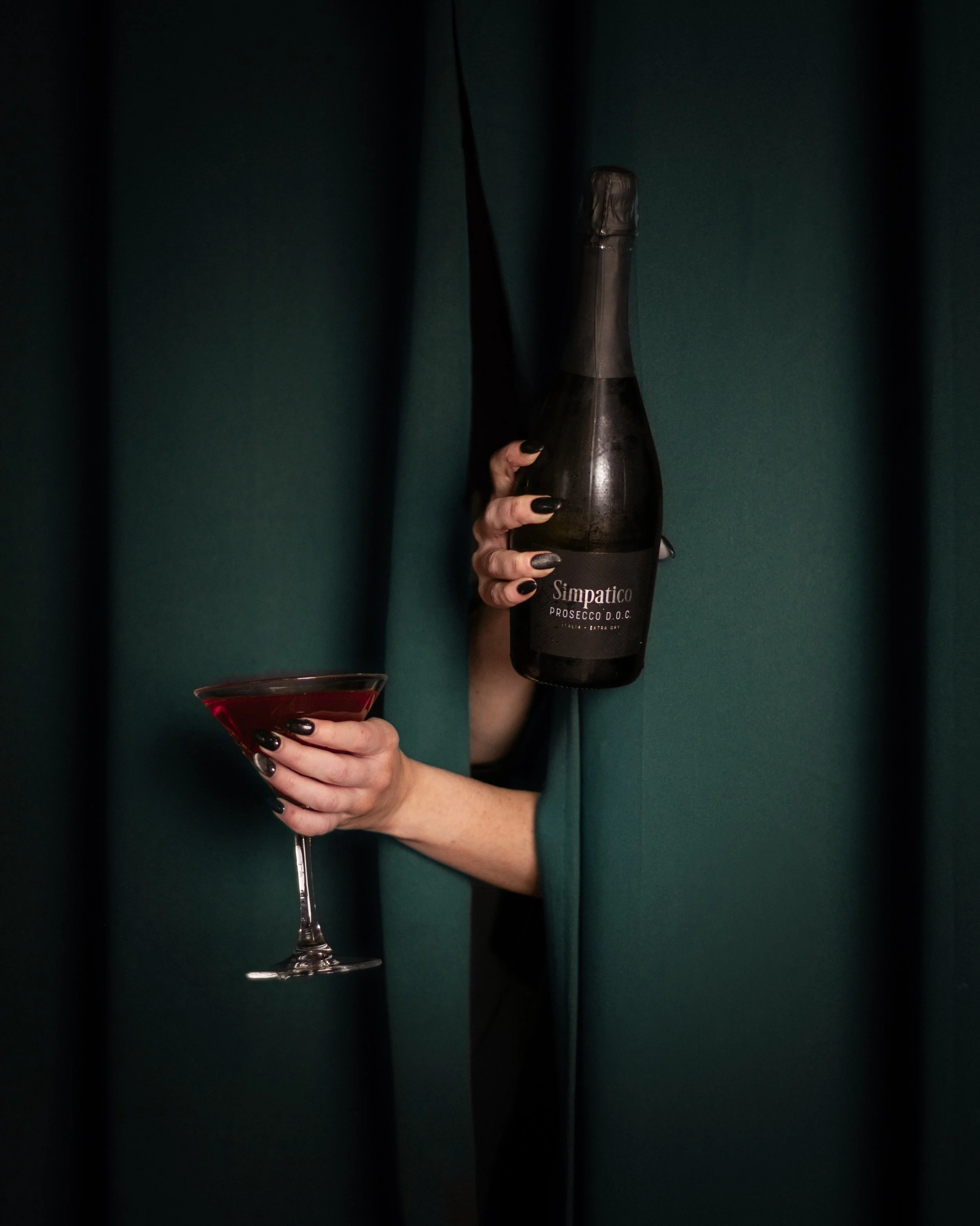 Two hands are emerging from a dark blue/green curtain, one hand holds a bottle of prosecco above, the other hand hold a martini glass full of red liquid.