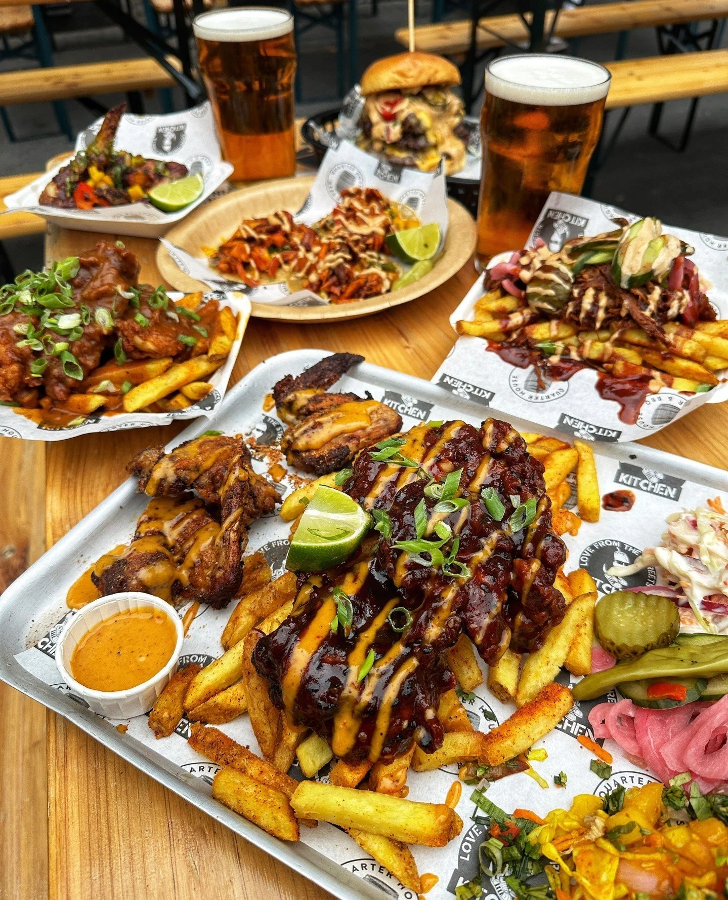 A wooden table outside full of food. In the fore front is a tray of BBQ food containing wings, thighs, chips, salad and slaw. In the background there are burgers, tacos, loaded fries and 2 pints of beer on the table.