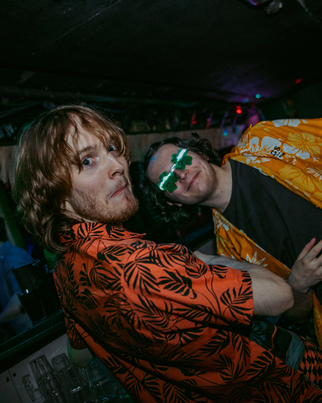 Two bartenders in tiki shirts and looking at the camera, the one closest is pulling a funny face with wide eyes and a downturned smile. The one behind him smirks with glasses on.