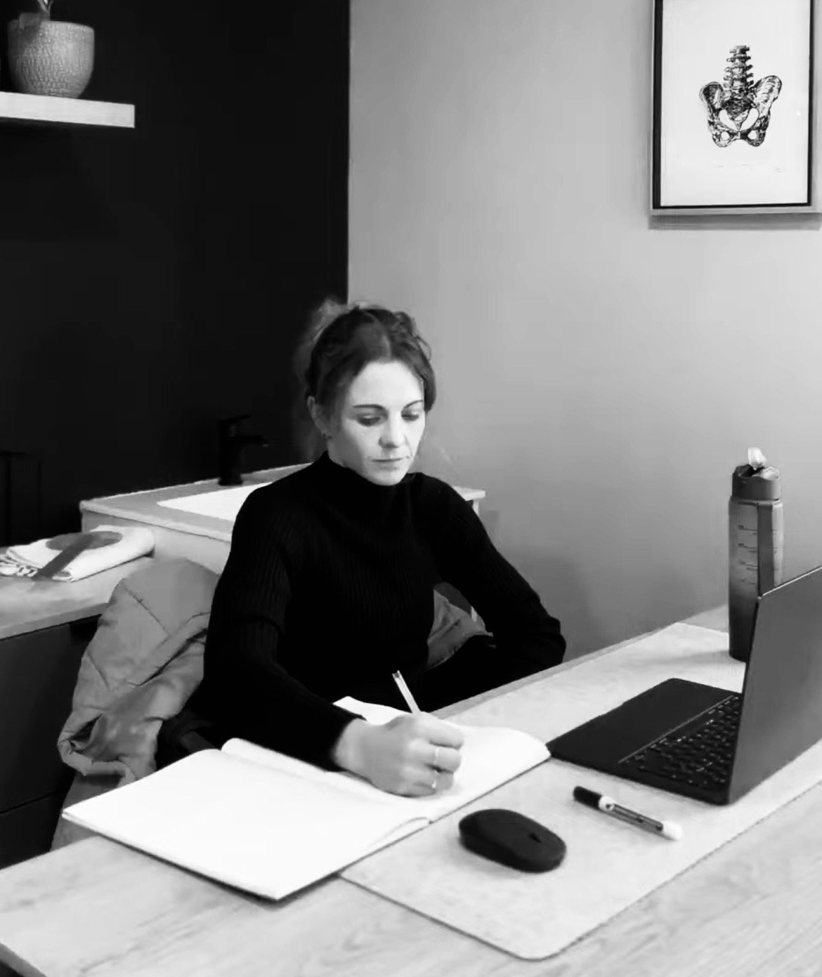 A woman with dark hair in a bun sits at a desk, writing in a notebook, with a laptop, mouse, and water bottle on the desk, in a room with framed artwork on the wall.