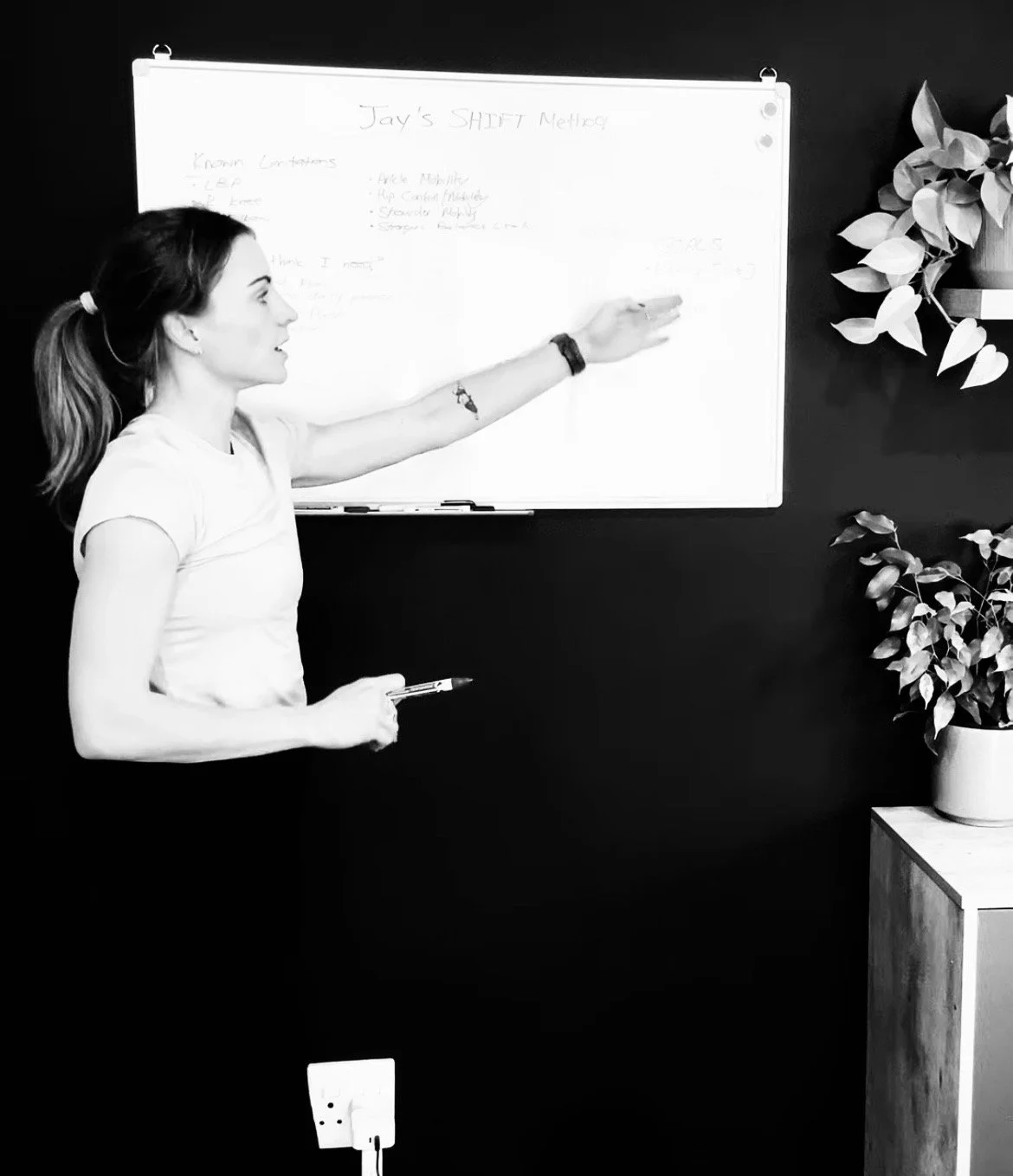 A woman standing in front of a whiteboard, pointing at it with her left hand while holding a marker in her right hand. The whiteboard has handwritten notes titled "Jay's SHIFT Methods". There is a potted plant on a wooden stand to the right of her, against a dark wall.