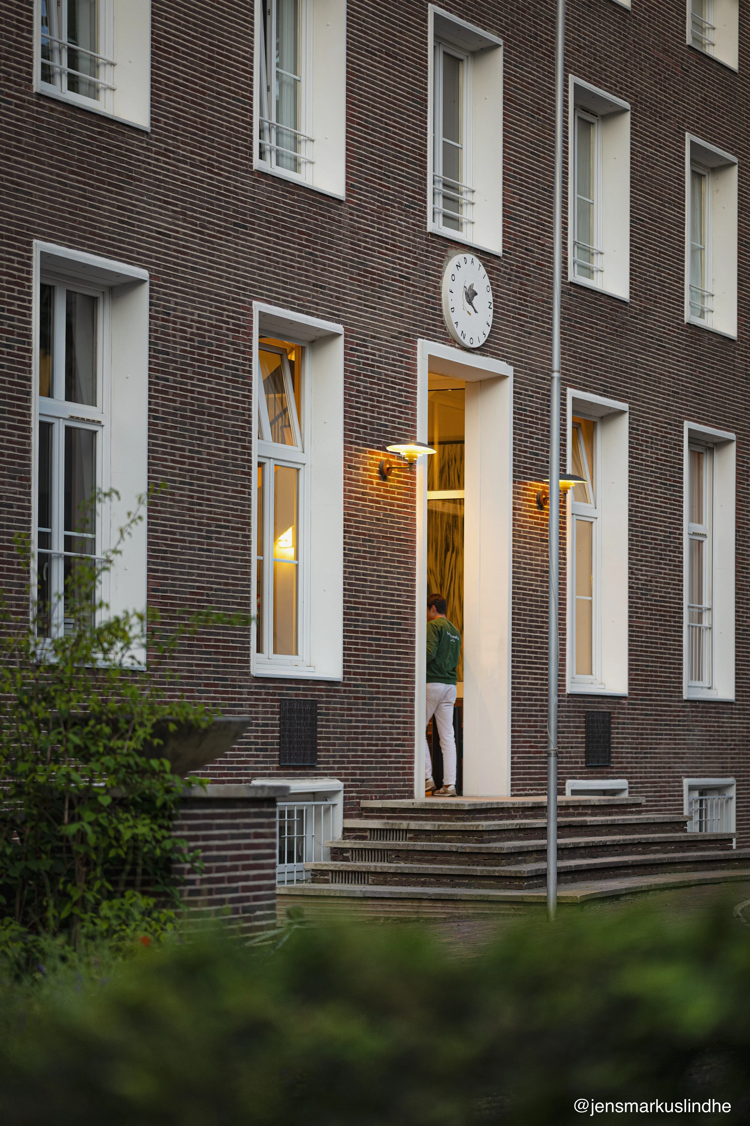 En rødbrun murstensbygning med flere hvide vinduer, nogle med åbne vinduesrammer. En person står i en åben skydedør, og der er udendørs væglamper, der lyser. Der er trappe foran bygningen og en facade med et ur, der viser tidspunktet, samt et skilt med teksten 'FOND A'.