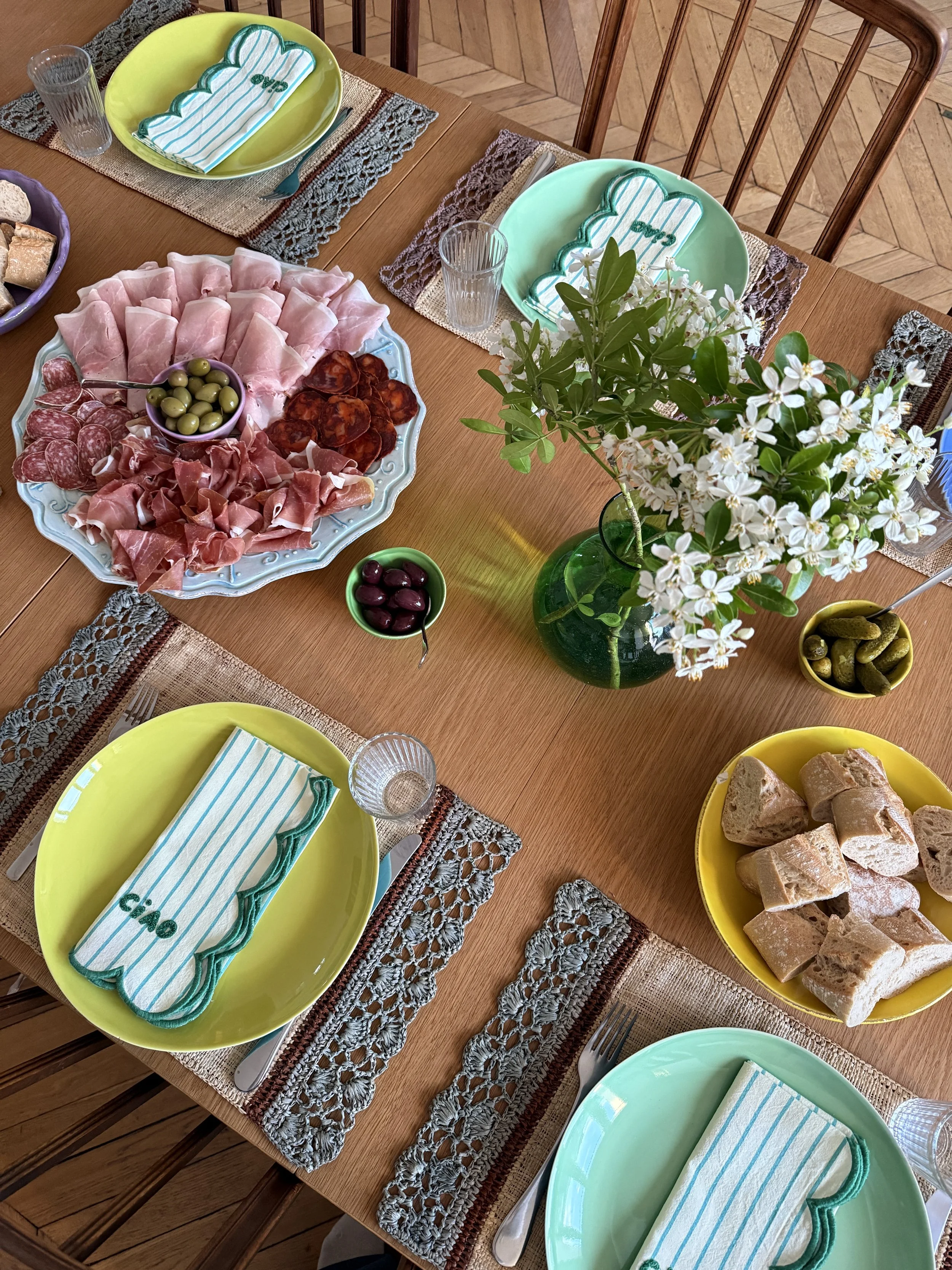 Et middagsbord dækket med farvede tallerkener, bestik, glas, salatbestik, en kop, en stor serveringsplatte med forskellige skinker, en skål med oliven, en vase med hvide blomster, og forskellige typer brød på en træbord med broderede underlag.