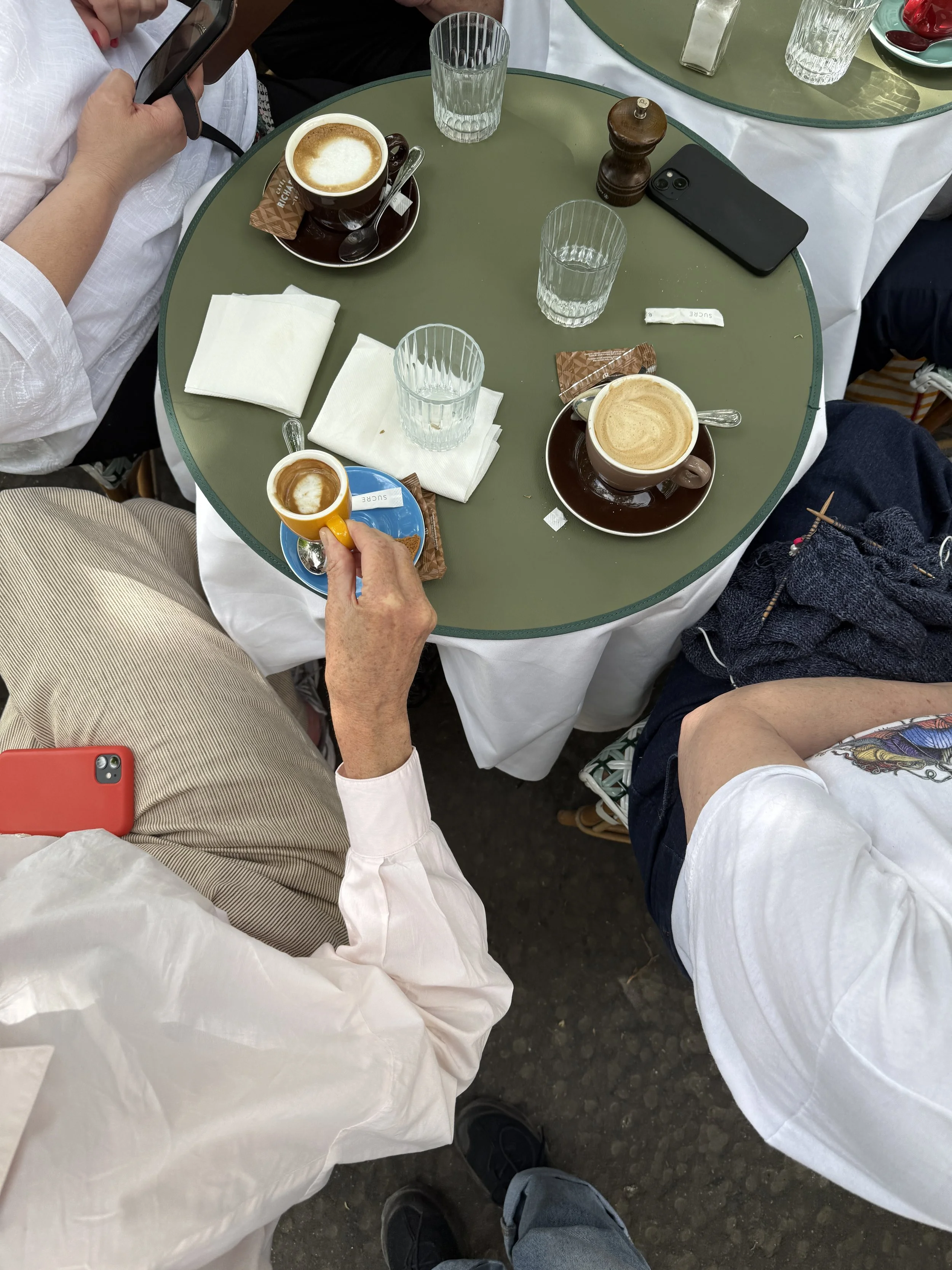 Billedet viser et bord med tre kopper kaffe med mælk eller cappuccino, nogle glas med vand, en krydderikværn, en seddel, papirlommetørklæder og en smartphone. Fire personer sidder omkring bordet, hvoraf to er synlige, og deres ben kan ses. Bordets overflade er grøn, og der er en hvid dug under nogle af genstandene.