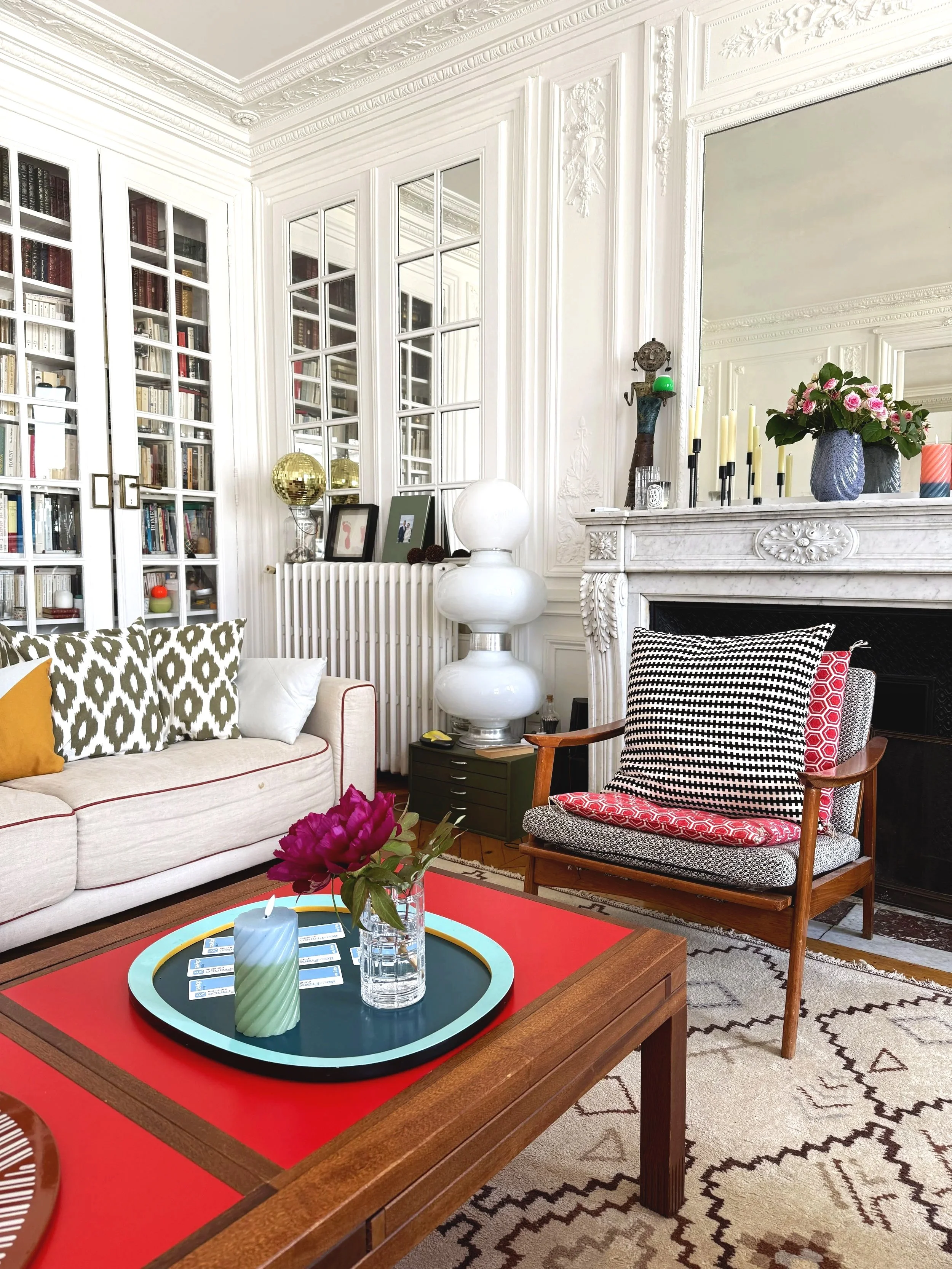 Et elegant stueindretning med en hvid sofa med pyntepuder i sort-hvid mønster, en træstol med en sort-hvid pude, en hvid marmorfunktion over en pejs, en rød sofabord med en grøn blomst i en vase, en sort rund bakke med lysestager og pynt, en stor spejlrefleks over pejsen, hvide vinduer med rammer, og dekorative elementer som guldballoner, billeder, og en lysestage.
