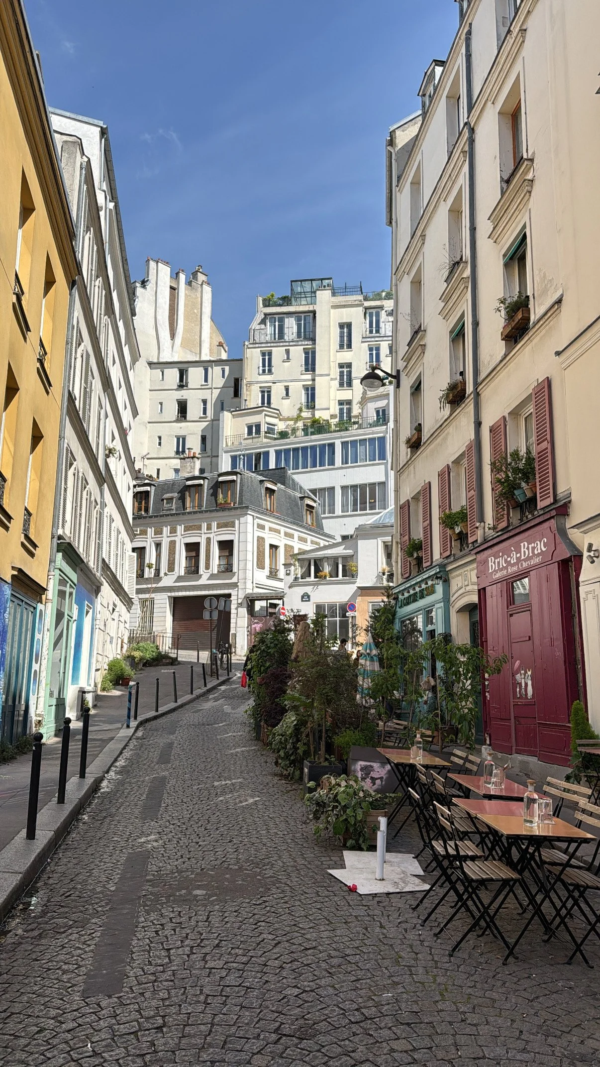 En hyggelig gade med cafeer med borde og stole udenfor, omgivet af gamle bygninger med blå, gule og cremefarvede facader i Paris.