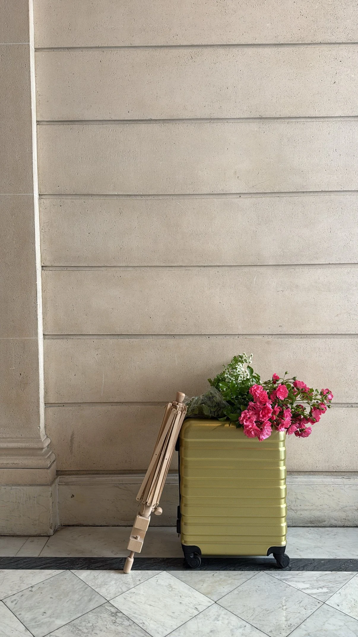 En grøn kuffert med fire hjul, en plante med pink blomster, en blomsterpotte med hvide blomster, og en sofa i beige.