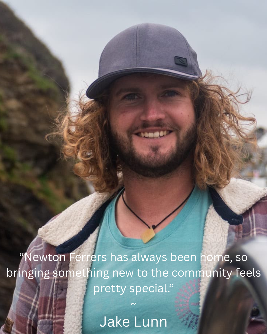 A smiling man with curly hair wearing a gray cap, teal shirt, layered jacket, and a necklace. In the background, there are rocks and an outdoor setting.