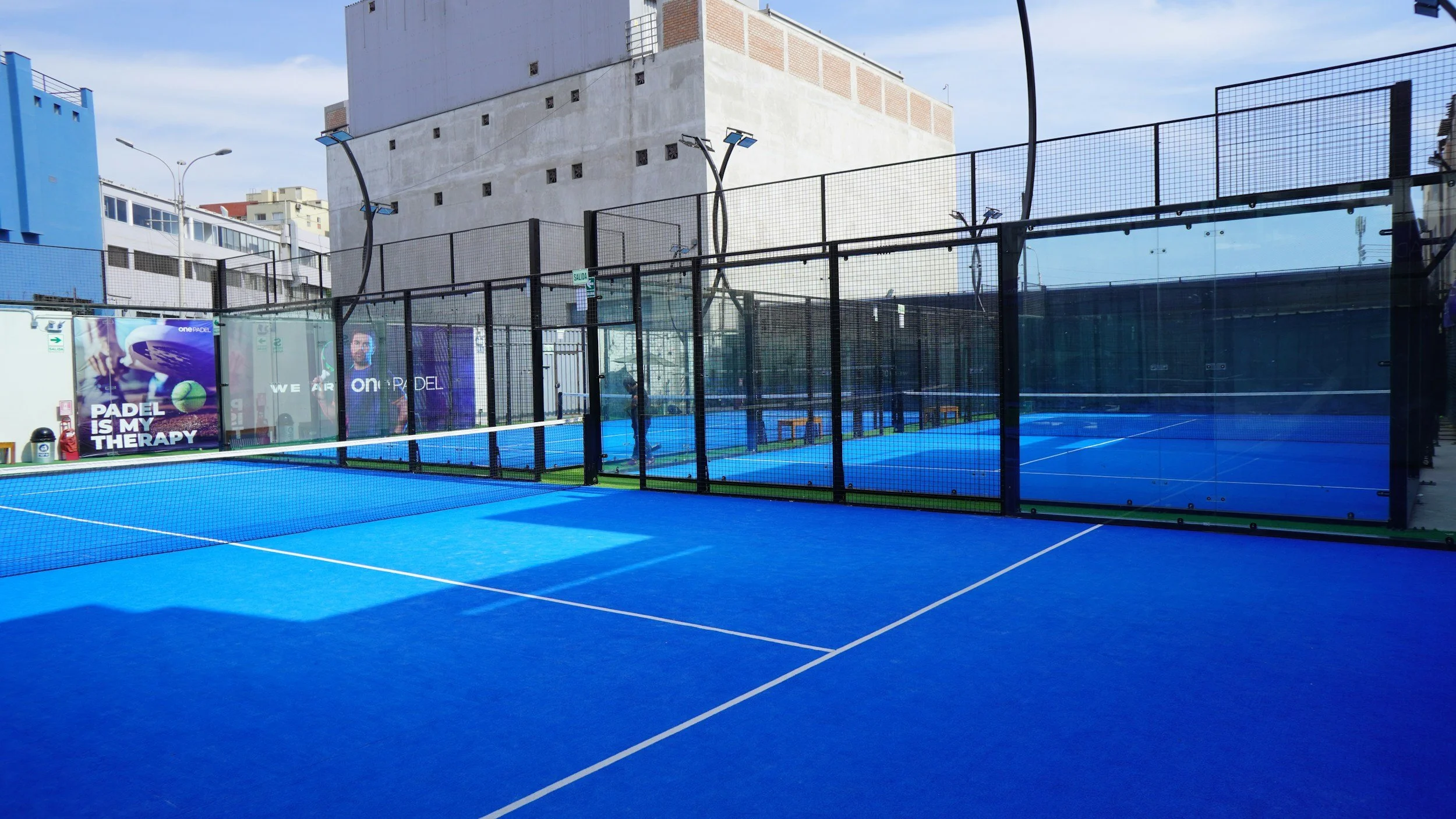 Outdoor padel tennis court with blue surface, enclosed by black glass and wire fencing, in an urban area with surrounding buildings and advertisements, sunny sky.