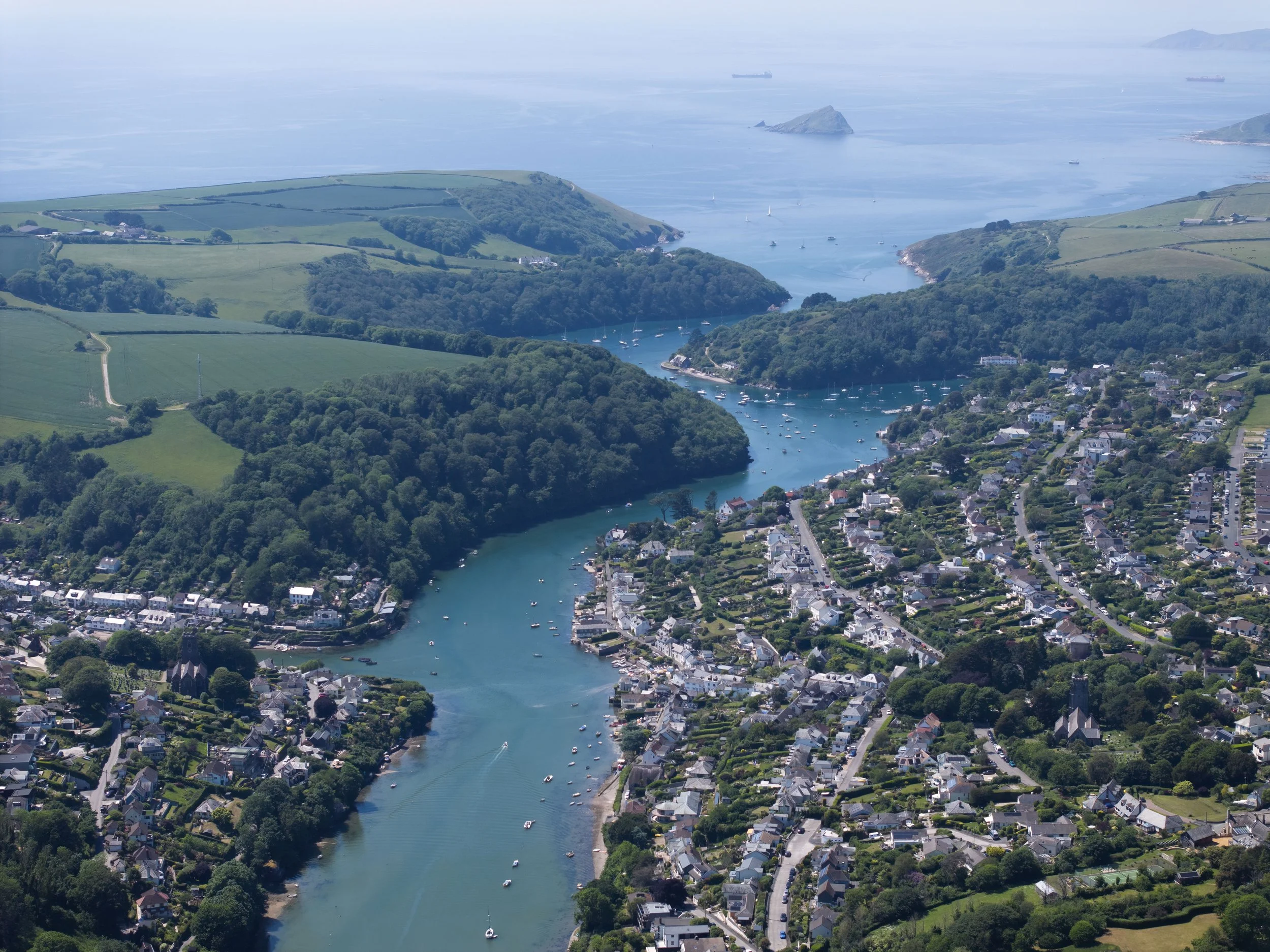 Aerial view of a coastal town with a river flowing into the ocean, surrounded by green hills and sailboats in the water.