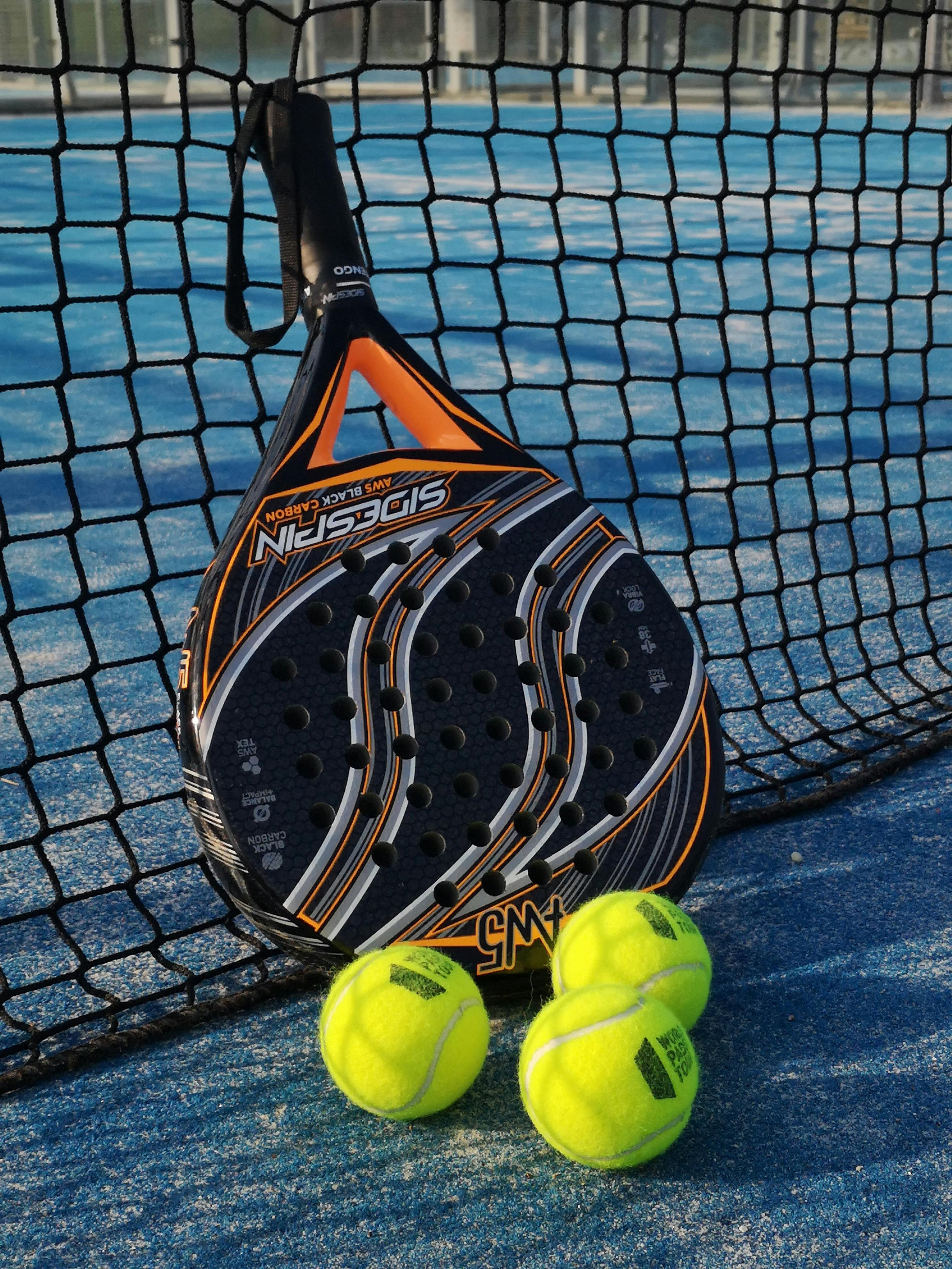Padel racket leaning against a net with three tennis balls on the court.