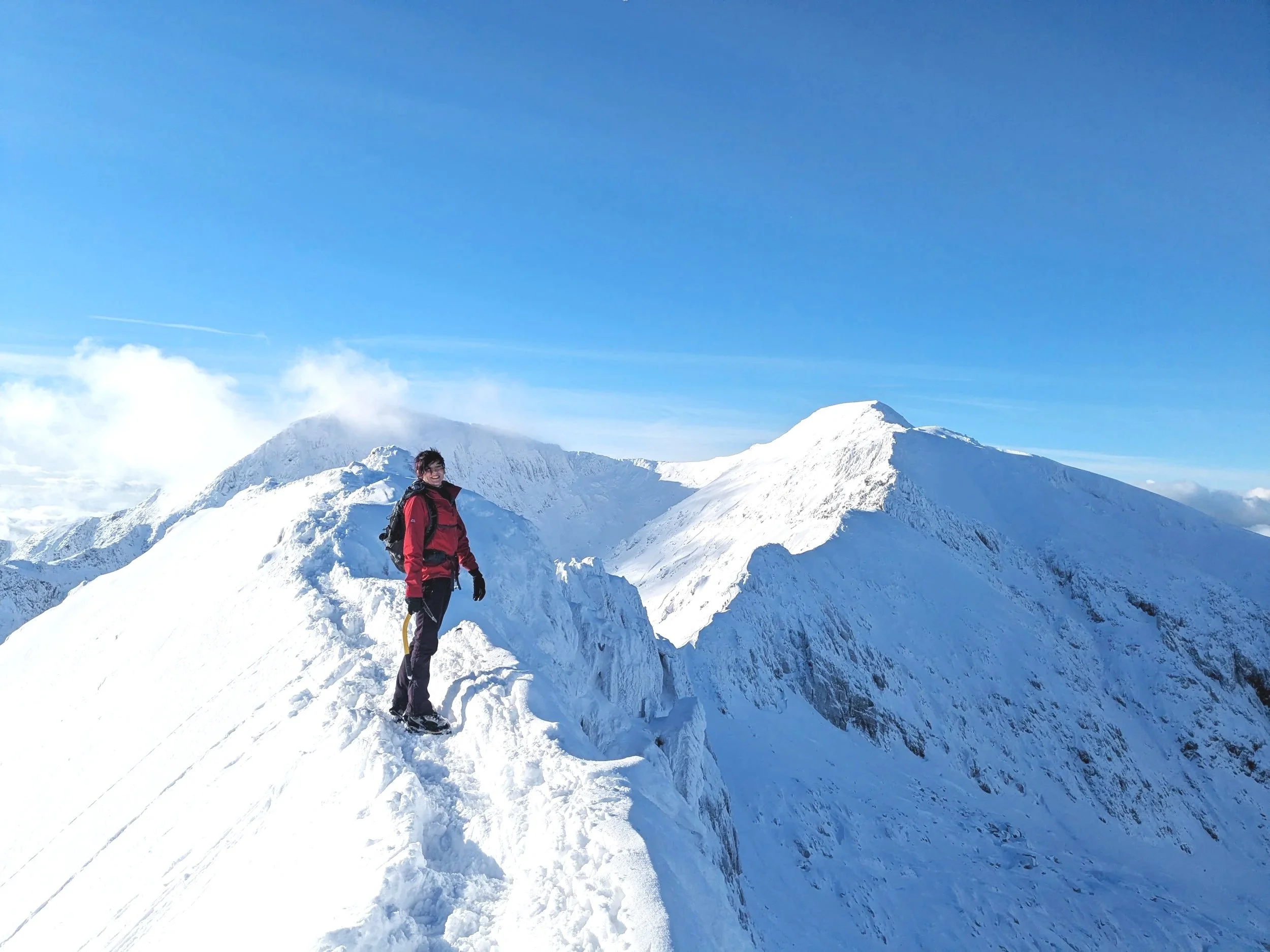 A person in a red jacket and black pants standing on a snowy mountain ridge with snow-covered peaks and a clear blue sky in the background.