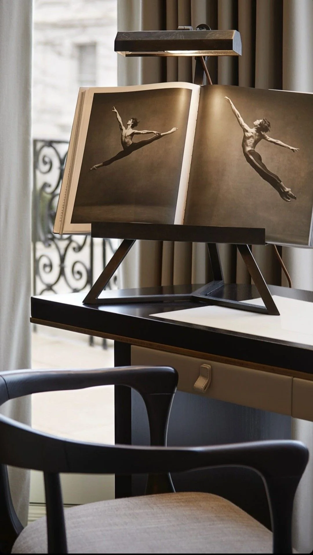 Et bokstativ med en åpen bok som viser svart-hvitt bilder av en ballettdanser i ulike hopp. Bokstativet plassert på et skrivebord i et rom med gardiner og vindu.