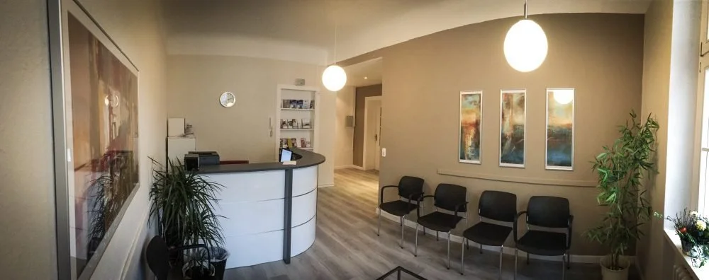 Wartezimmer mit fünf schwarzen Stühlen, Pflanzen, Wandkunst und Empfangstresen in heller Einrichtung.