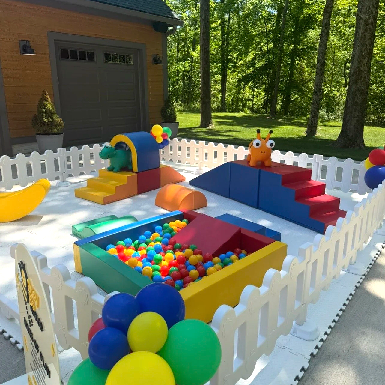 Colorful outdoor children's play area with ball pit, slide, and foam climbing structures, surrounded by white picket fence and set in a backyard with trees.