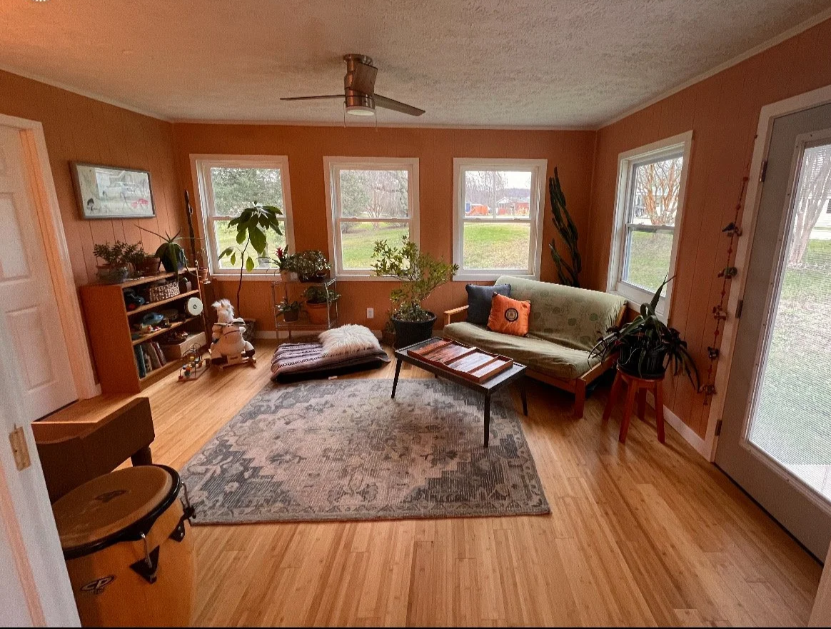 A bright living room with tan walls, hardwood floors, and multiple windows. It contains a green sofa with pillows, a wooden coffee table, a bookshelf, potted plants, and a ceiling fan.