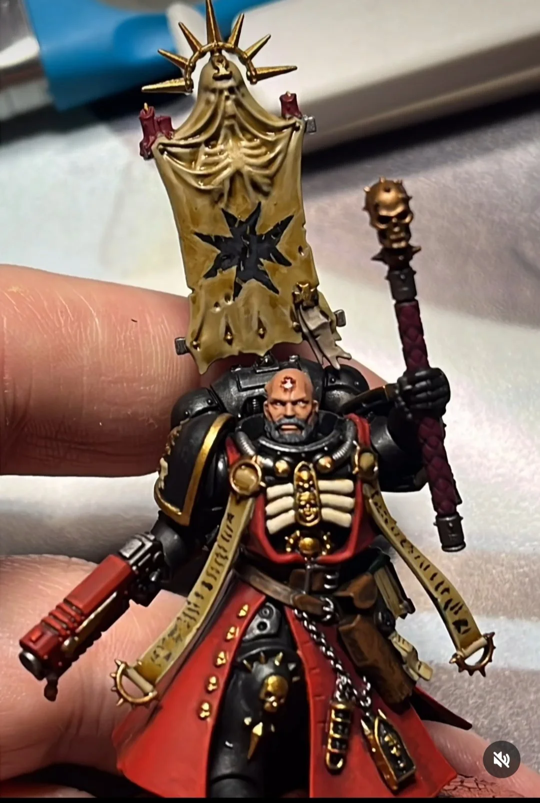 A detailed miniature figurine of a medieval warrior wearing black armor with gold accents, a red cape, and a skull motif. He holds a staff with a spiked metallic head in one hand and a firearm in the other. Behind him is a banner with black symbols o