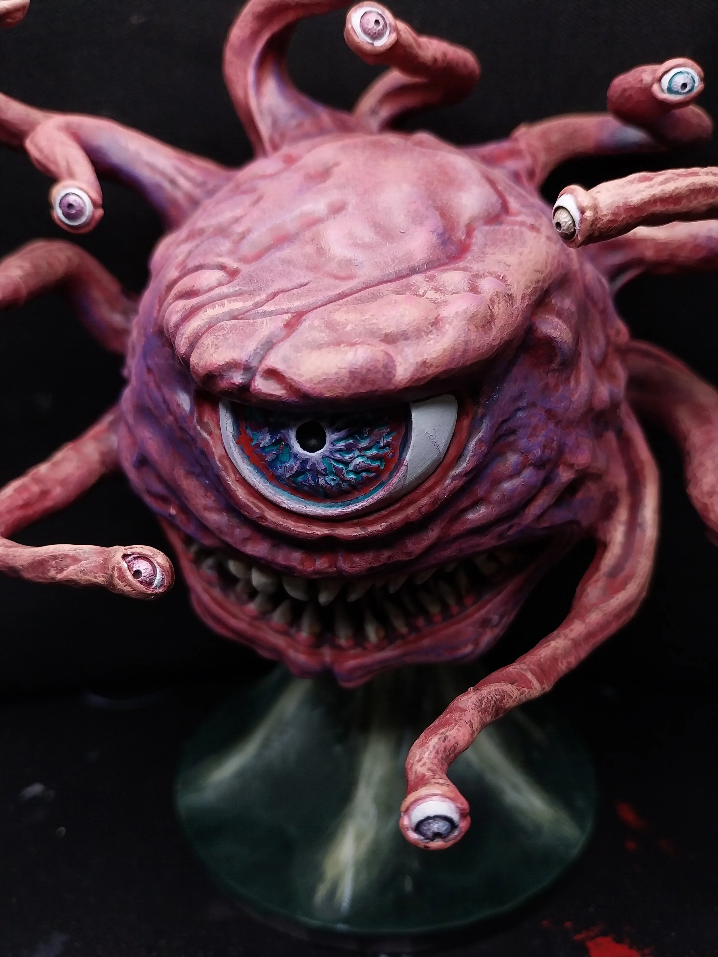 A surreal sculpture resembling a creature with a large, pinkish, textured head, multiple eye stalks with eyeballs, a single large eye with a colorful iris, and a wide, jagged mouth with sharp teeth, mounted on a green base.