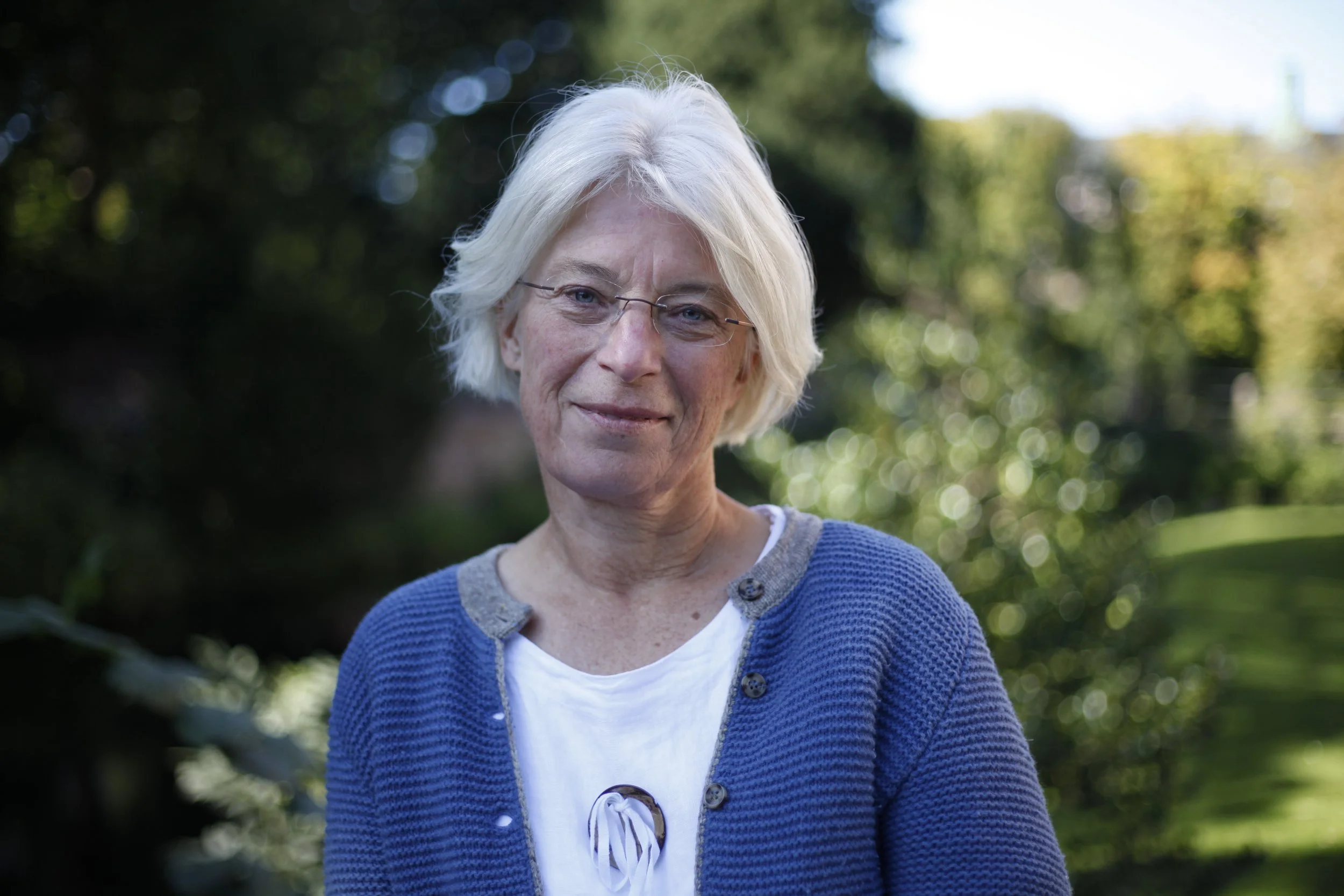 Ältere Frau mit weißen Haaren, trägt eine blaue Strickjacke und eine weiße Bluse, steht im Freien vor einem grünen, bewaldeten Hintergrund.