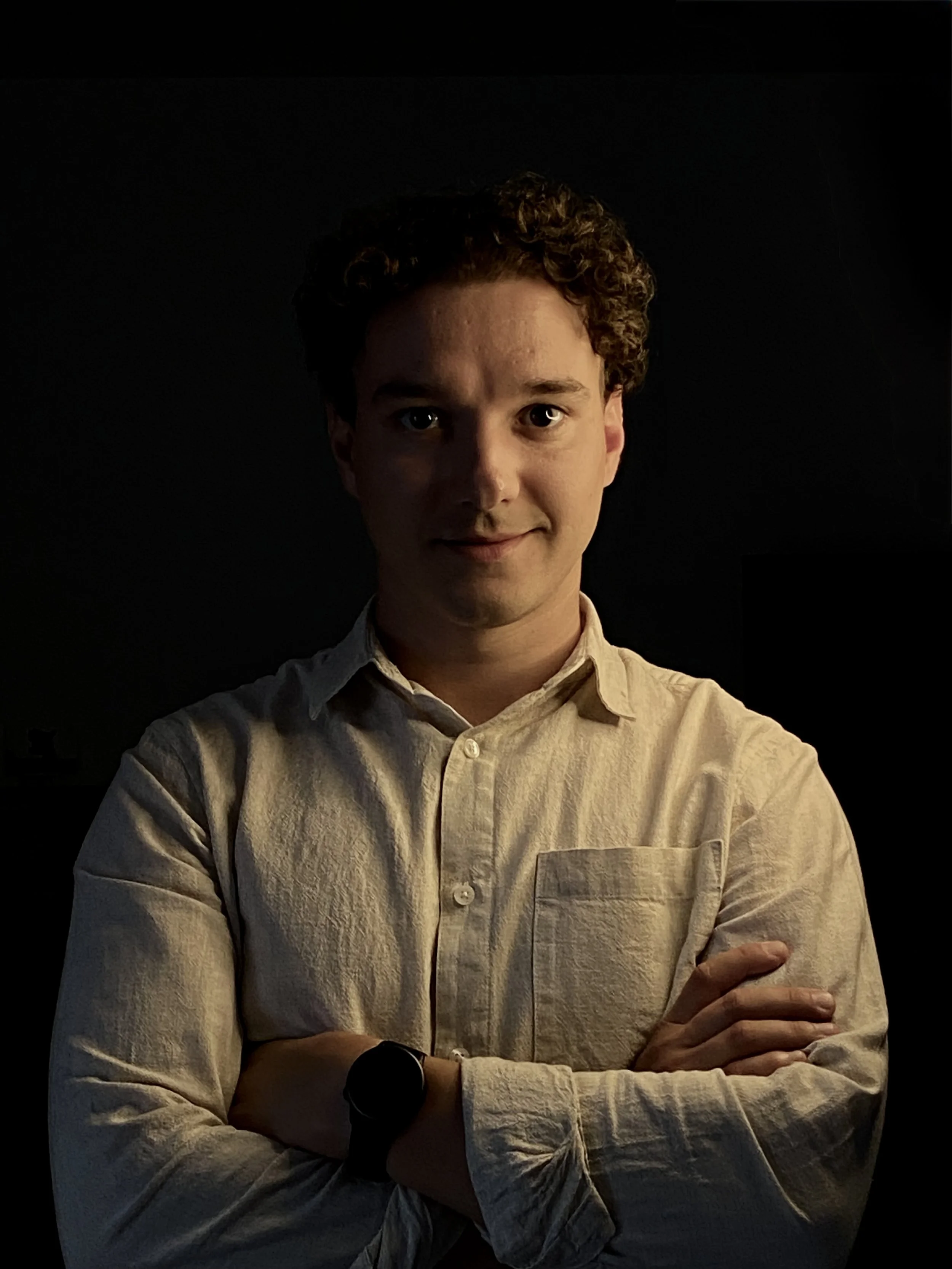 Portrait of a young man with curly hair wearing a light-colored shirt and a black smartwatch, crossing his arms, against a dark background.