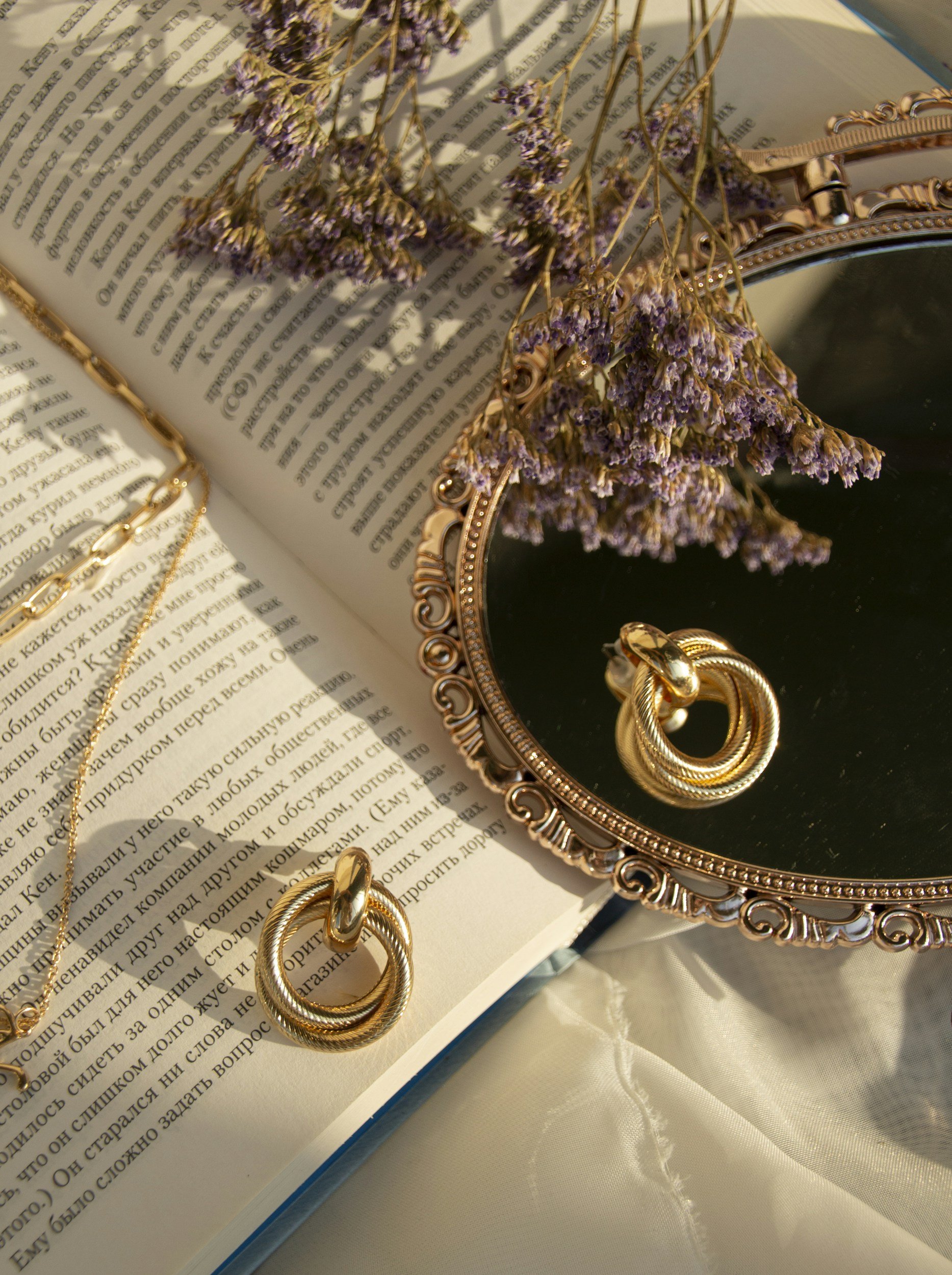 Jewelry collection with gold rings and necklace, purple dried flowers, and an ornate mirror on top of an open book with text, set on a white surface.