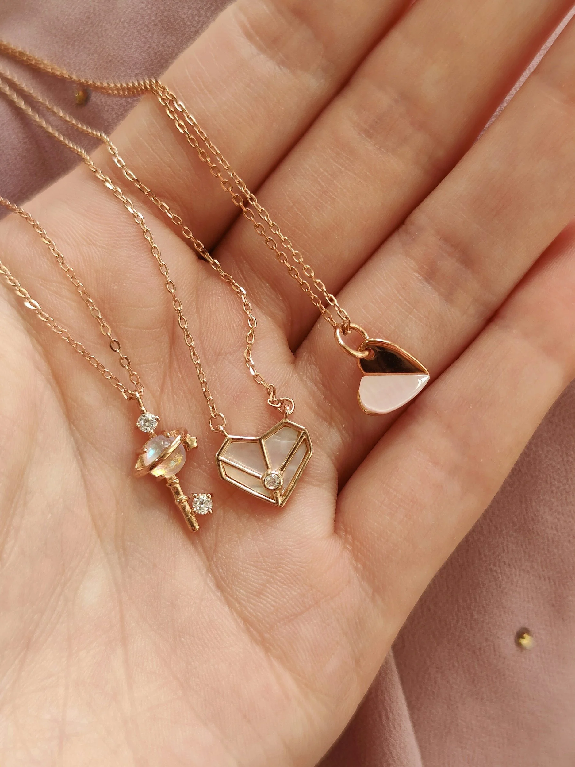 Three delicate gold necklaces with unique pendants: a key with small diamonds, a geometric stained glass heart, and a half-heart with a shiny gold finish, held above a pink fabric background.