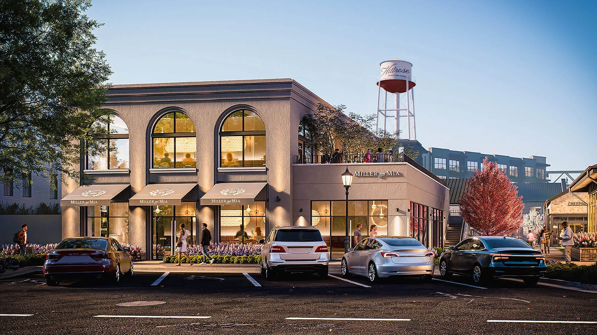A commercial building with a restaurant named Miller & MIA on the ground floor, featuring large arched windows and awnings. There are parked cars in front and people walking along the sidewalk. A tiered water tower with the word "Hilrose" is visible in the background. The scene is set during dusk or early evening, with trees and a clear sky.