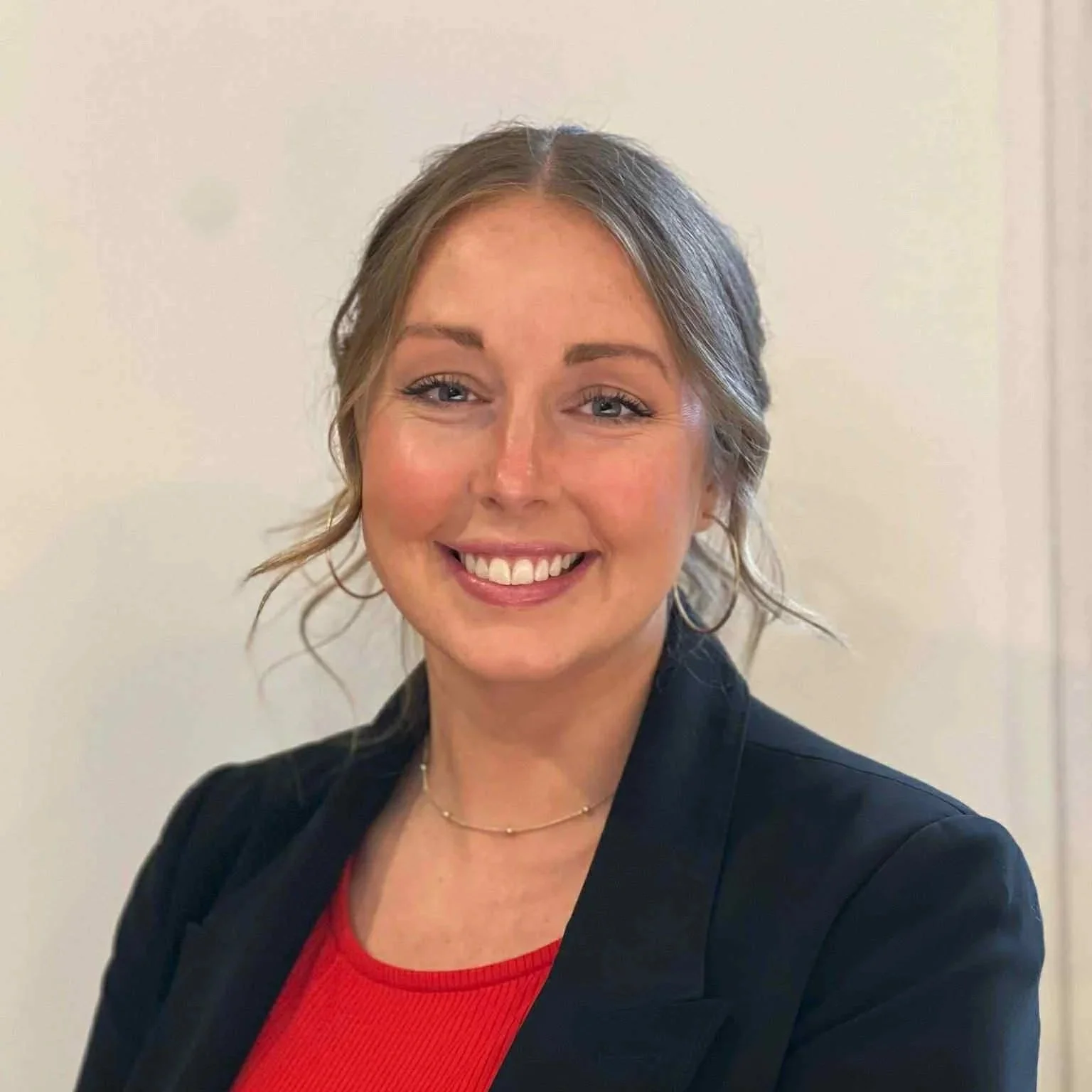 A woman with light skin, brown hair styled in loose waves, smiling with white teeth, wearing a black blazer, red top, and a delicate gold necklace, standing against a plain background.