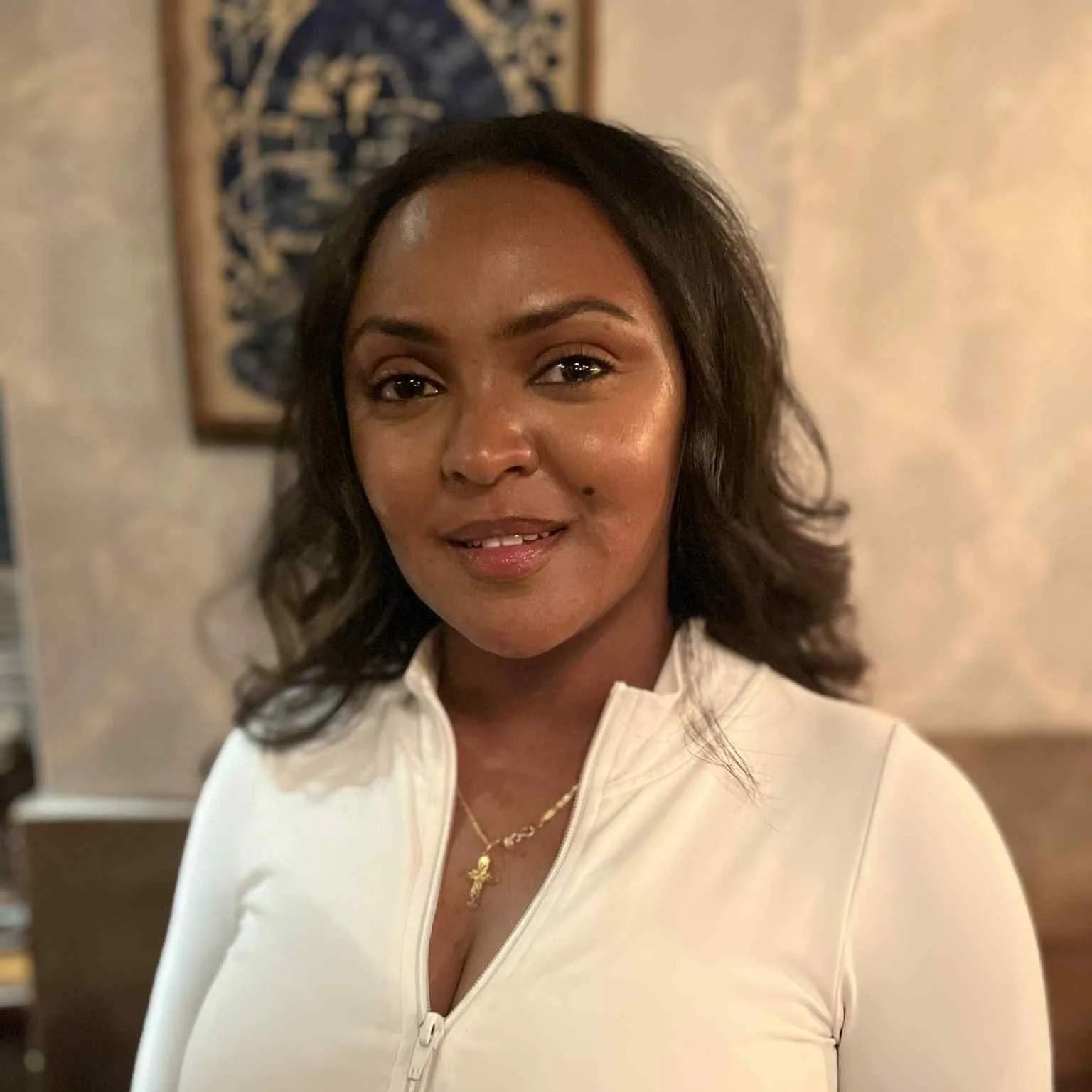 A woman with dark brown, wavy hair and warm brown skin, smiling at the camera. She is wearing a white zip-up top and a necklace with a small cross pendant. The background includes a blurred wall with a decorative art piece.