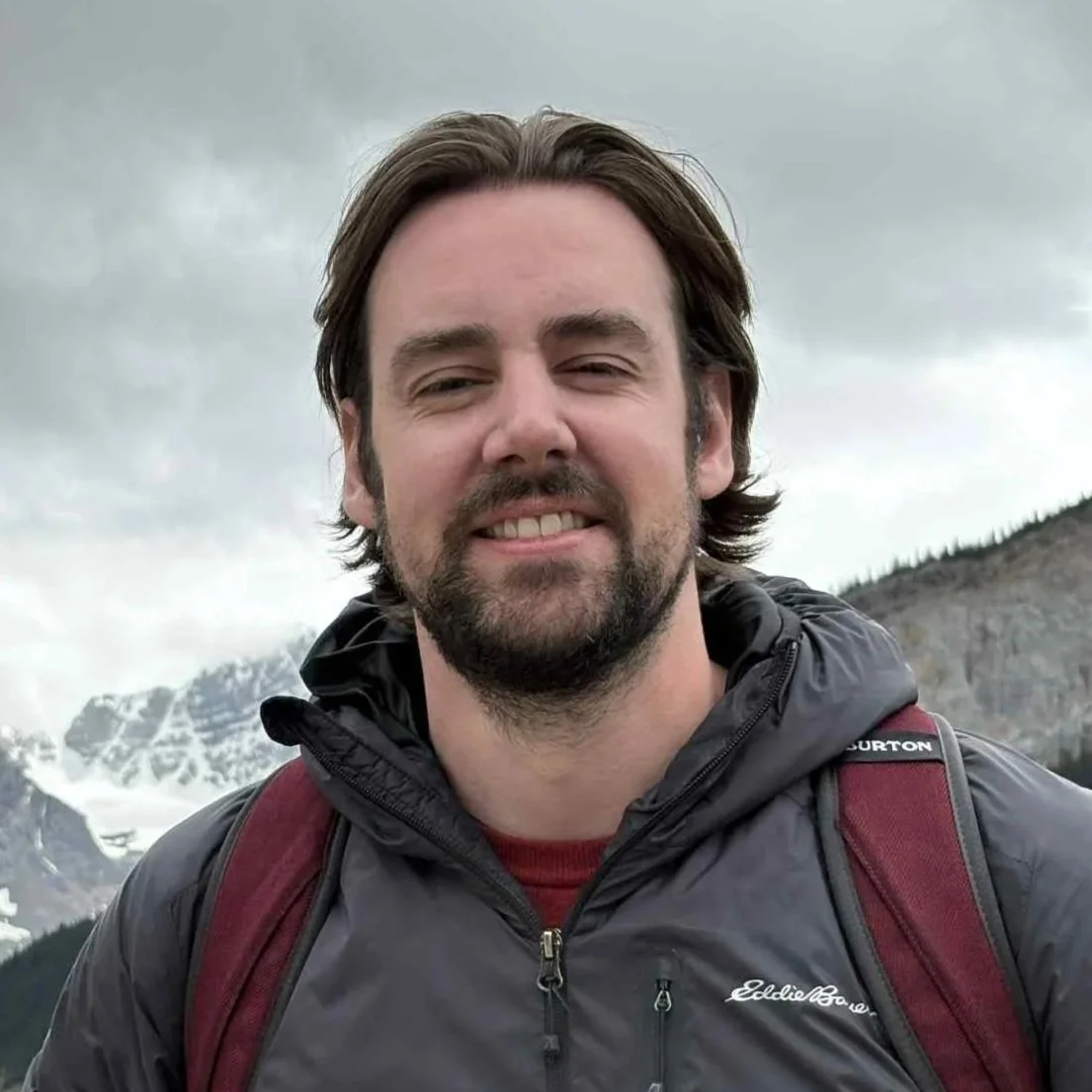 A man with dark hair and a beard smiling outdoors on a cloudy day, wearing a black jacket and red backpack, with mountains in the background.