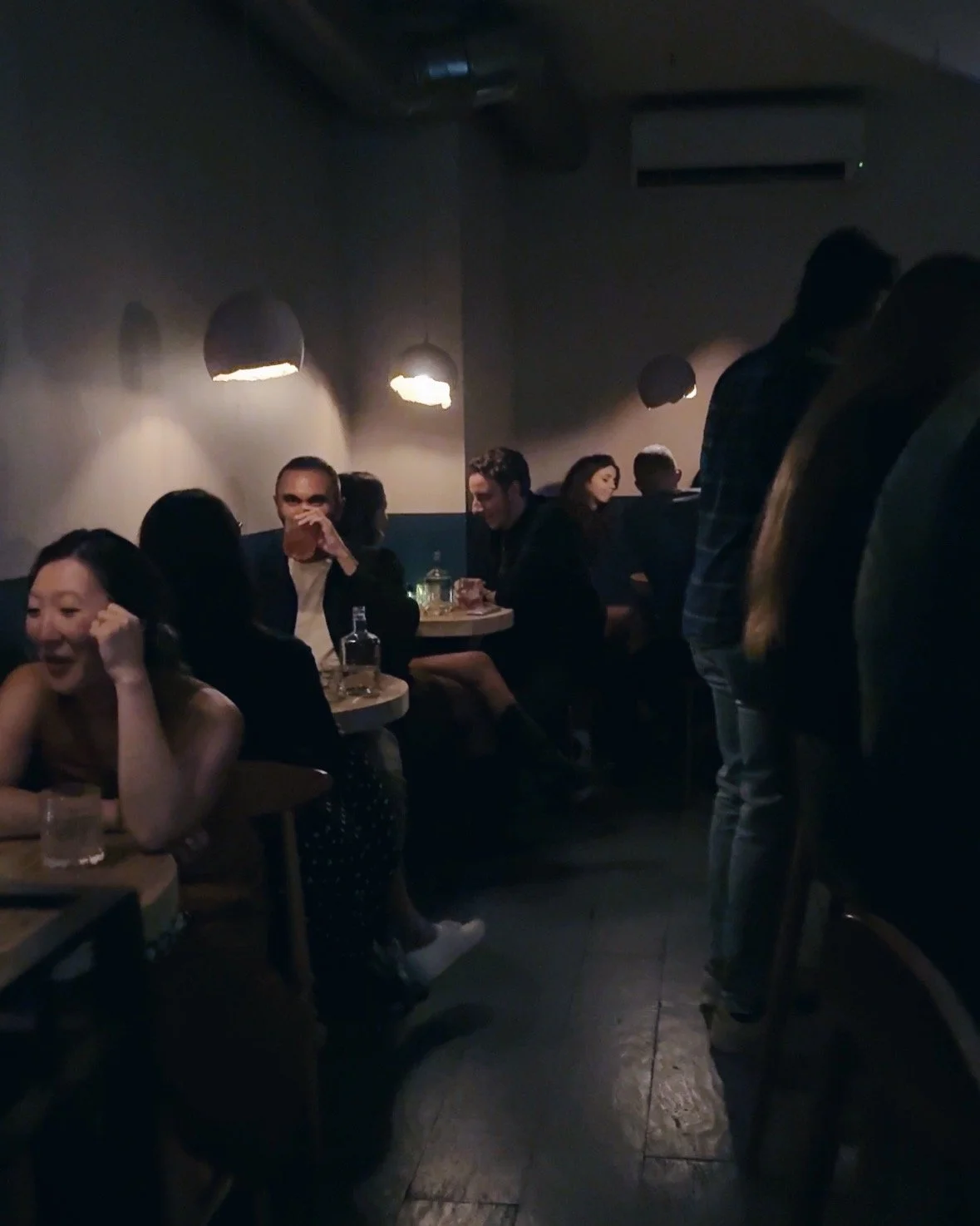 A group of bar patrons seated at small round wooden tables and gathered around the bar counter, enjoying drinks and engaging in conversation.