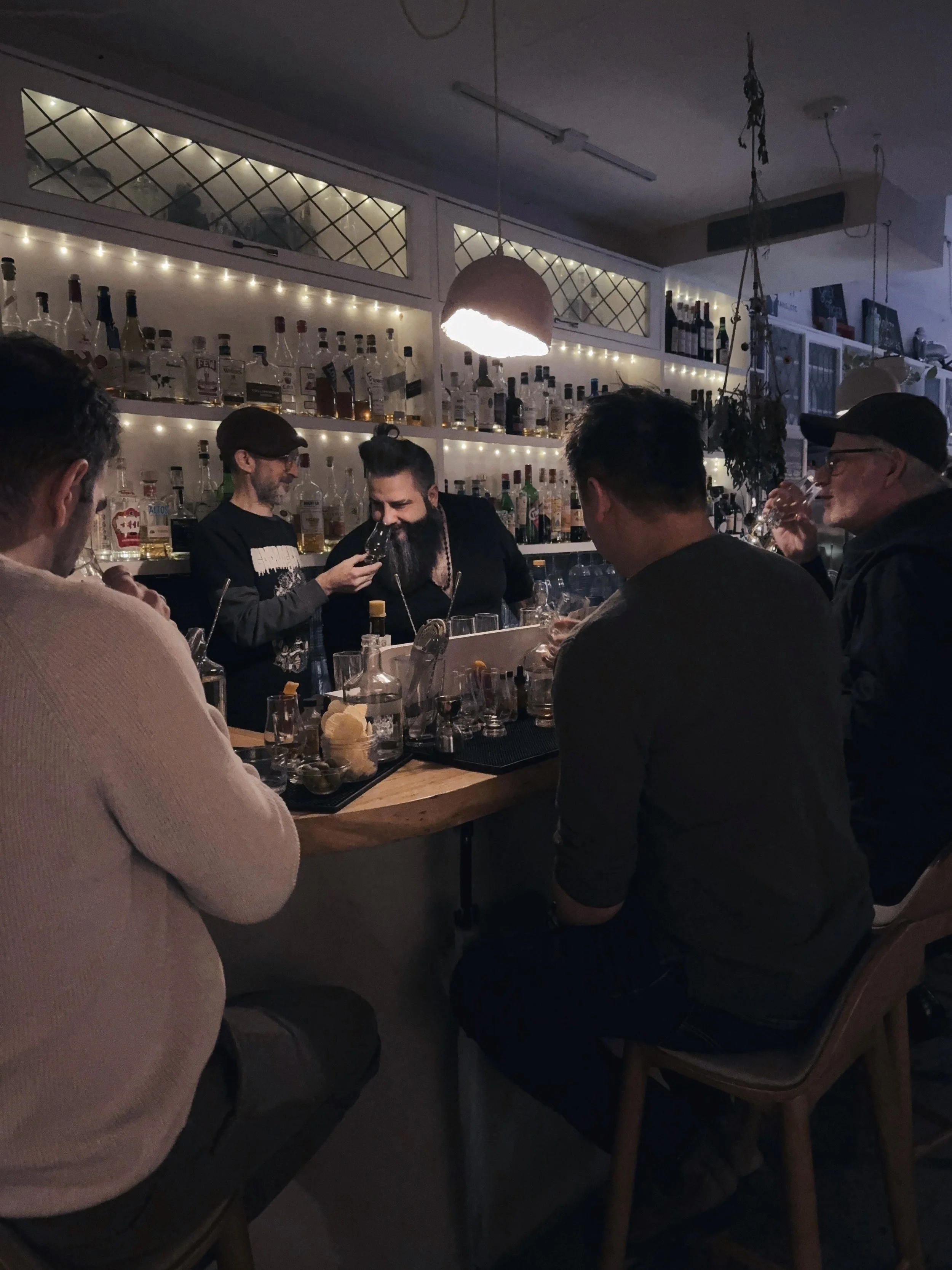 A group of people sitting at a bar counter in a dimly lit cocktail bar, with string lights and warm pendant lighting overhead, participating in a cocktail class while the instructor holds his Glencairn glass of whisky out for his assistant to nose.