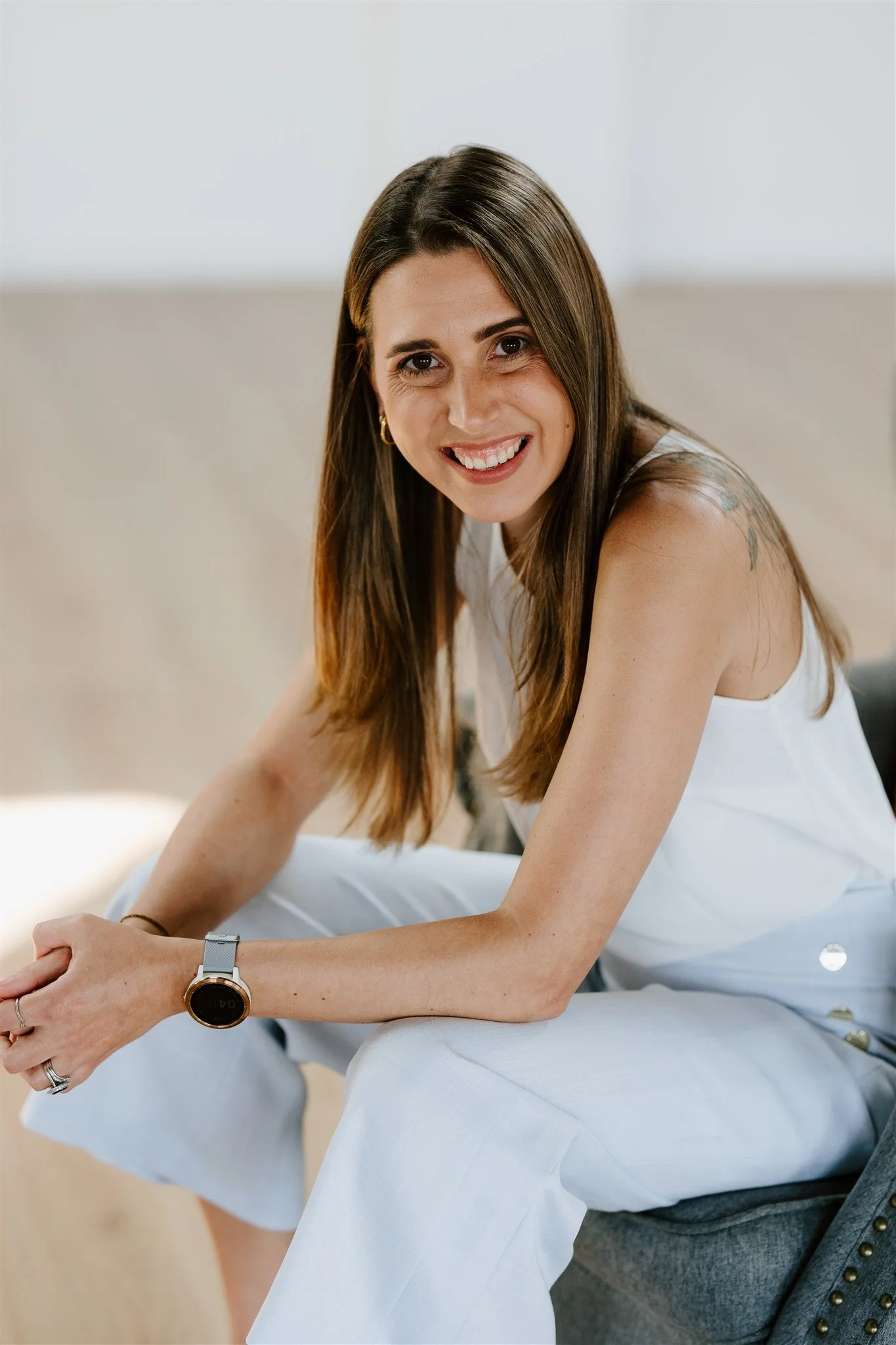 Close-up of a smiling woman with long brown hair, wearing a white sleeveless top, a gold hoop earring, and a watch, sitting on a light-colored surface.