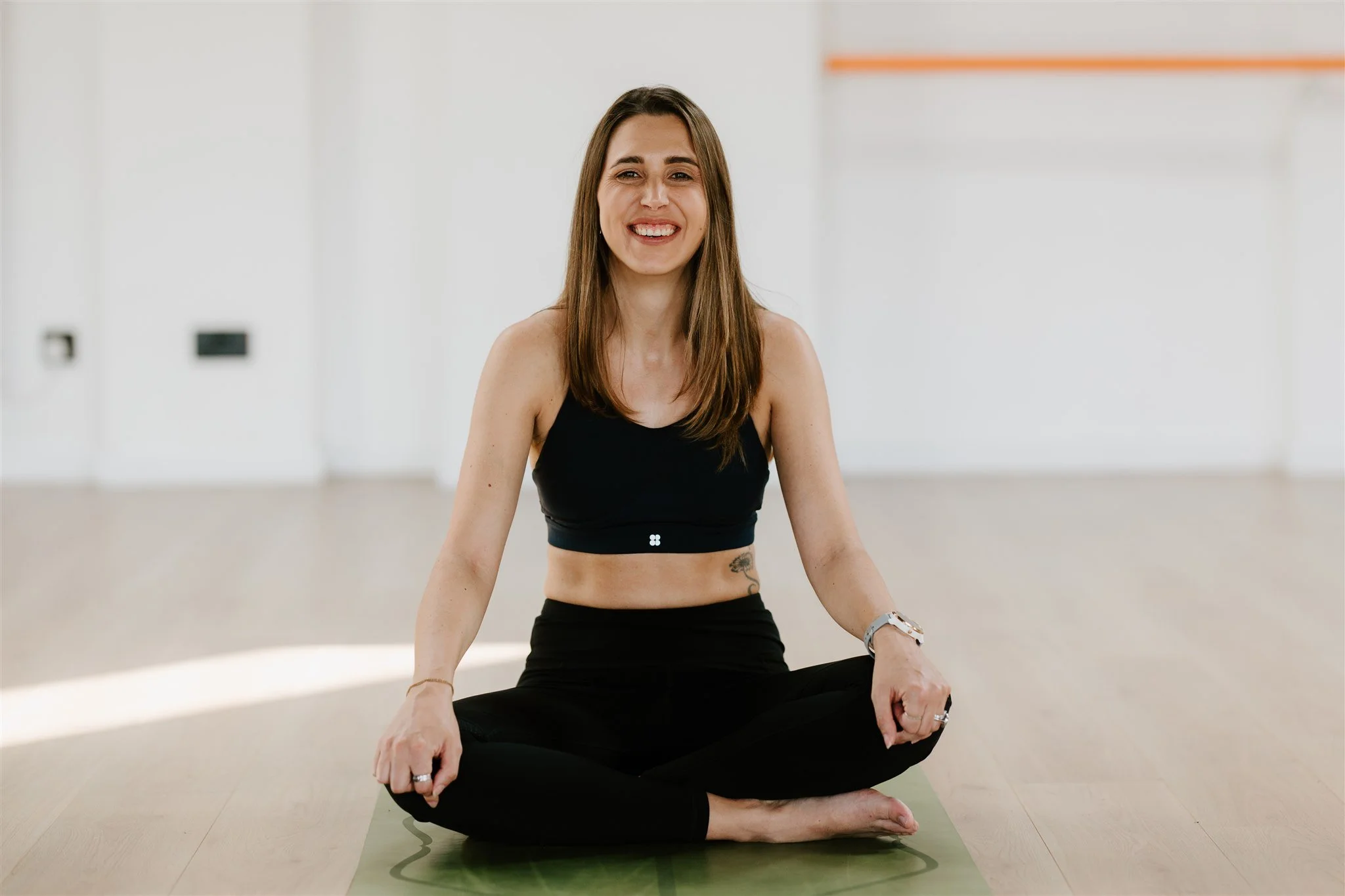 A woman practicing yoga in a dance studio, sitting cross-legged on a yoga mat, smiling at the camera.