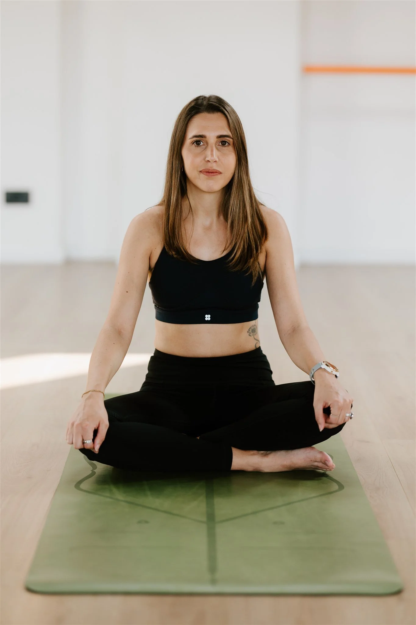 A woman with brown hair sitting cross-legged on a green yoga mat, practicing meditation in a bright, minimalistic room.