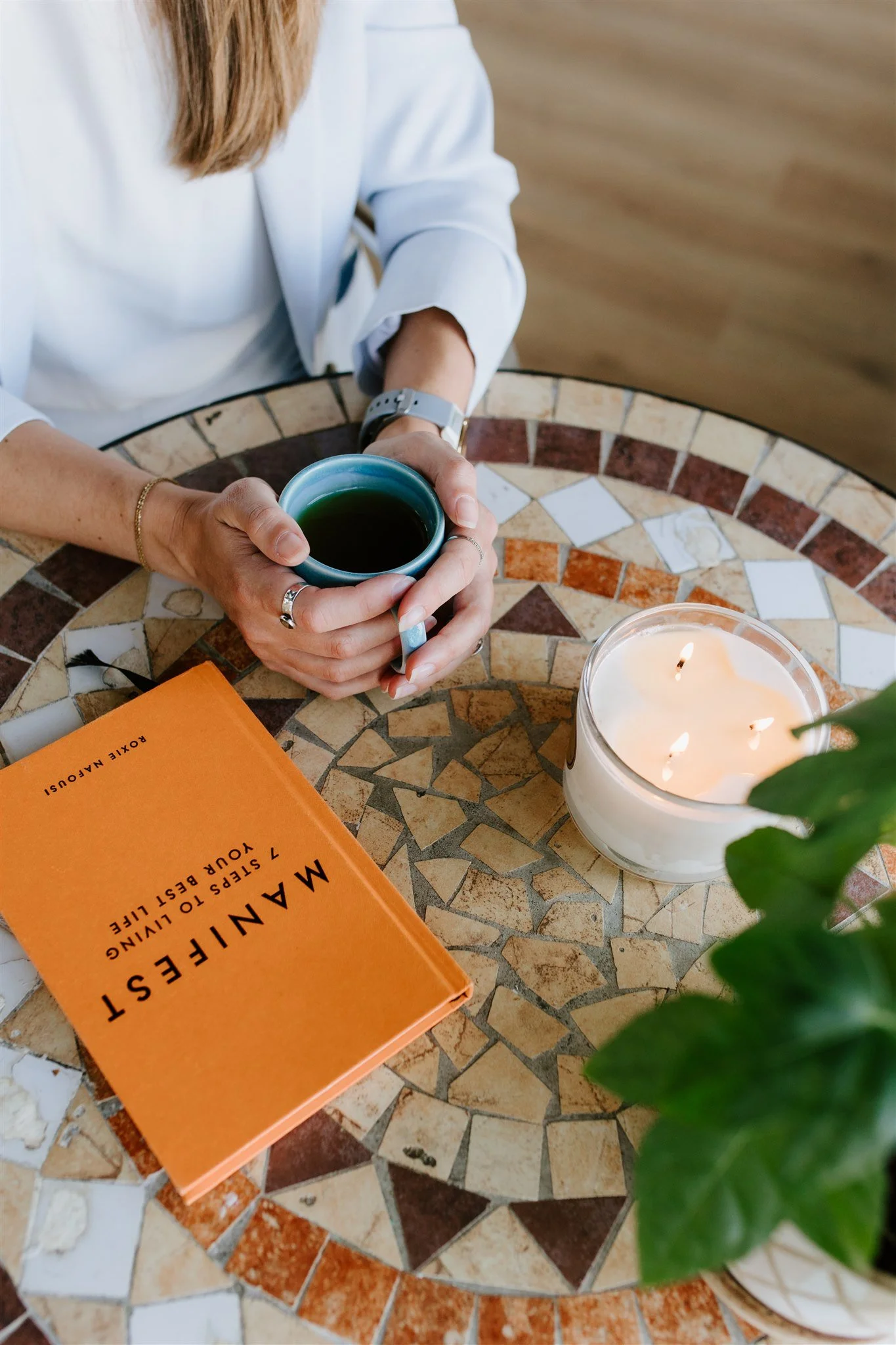 A person holding a dark-colored ceramic mug with a teal interior and black beverage inside, sitting at a round mosaic table with a lit white candle in a glass holder, an orange book labeled 'MANIFEST,' and a green plant partially visible. The person's hands are adorned with rings and bracelets, and they are wearing a white long sleeve shirt.