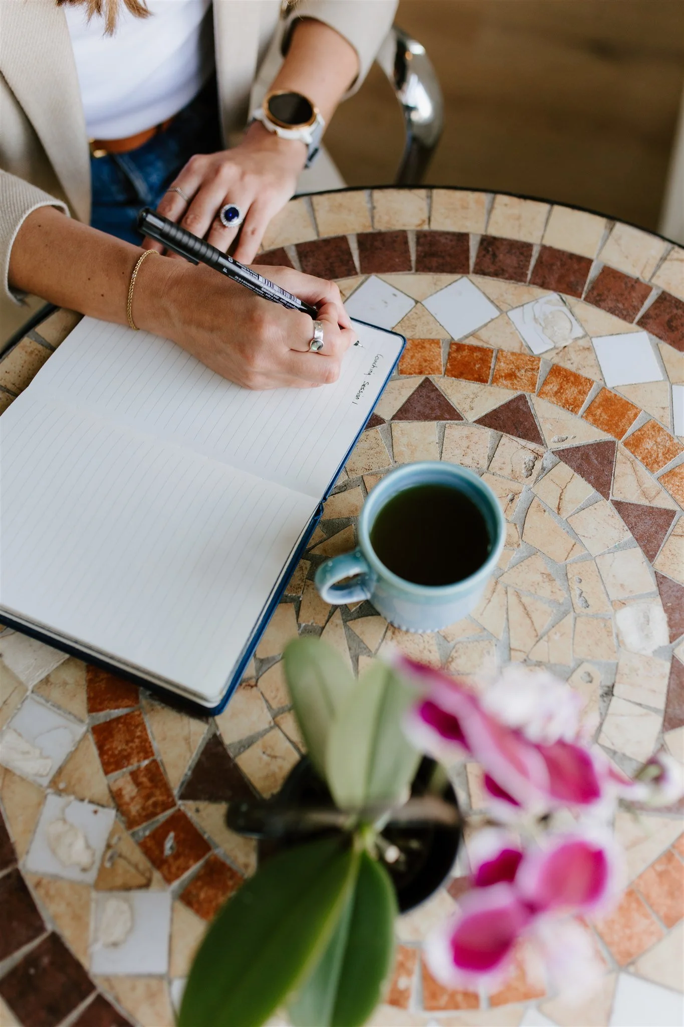 A person writes notes in a notebook on a mosaic-tiled table, with a cup of coffee and a pink orchid flower in the foreground.
