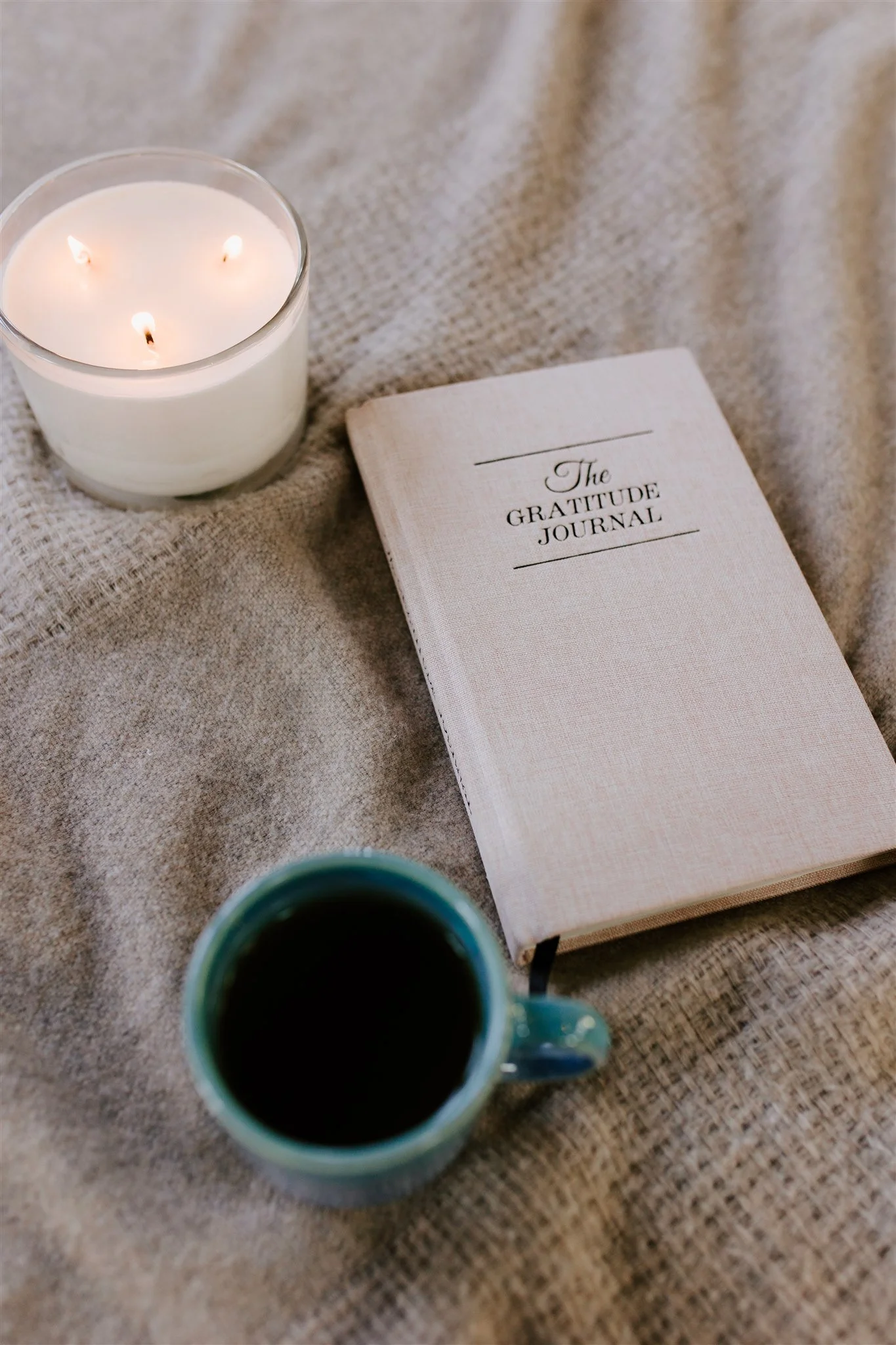 A lit white candle in a glass jar, a closed notebook titled 'The Gratitude Journal,' and a cup of black coffee on a beige textured fabric surface.