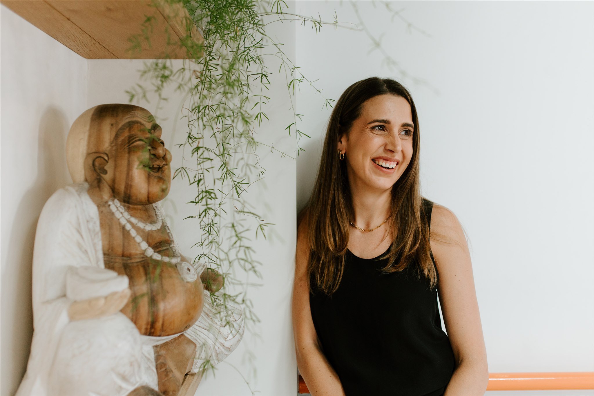 A woman with long brown hair, wearing a black sleeveless top, smiling while sitting next to a wooden sculpture of a smiling person, with green hanging plants above.