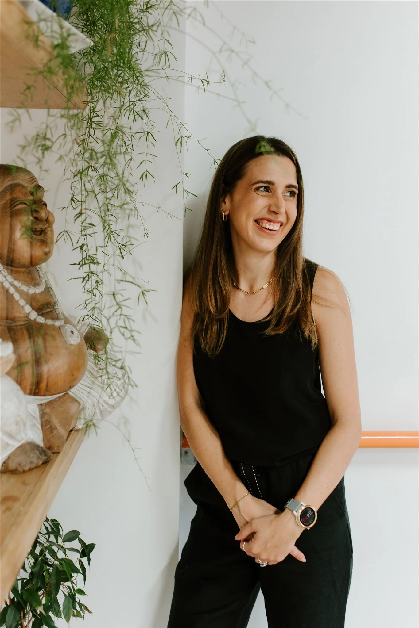 A woman with long brown hair wearing a black sleeveless top, gold jewelry, and a smartwatch, standing next to a wooden plant display with plants, smiling and looking to her right, against a white wall.