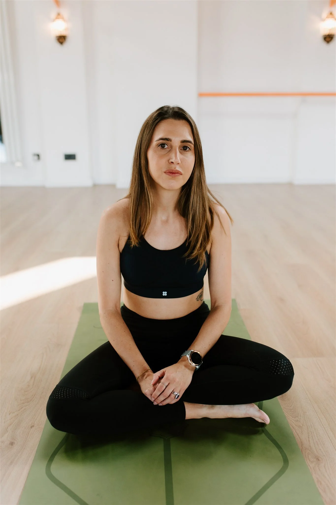 A woman sitting in a cross-legged yoga pose on a green yoga mat in a bright room, wearing black workout attire and a smartwatch, with her hands resting on her knees.