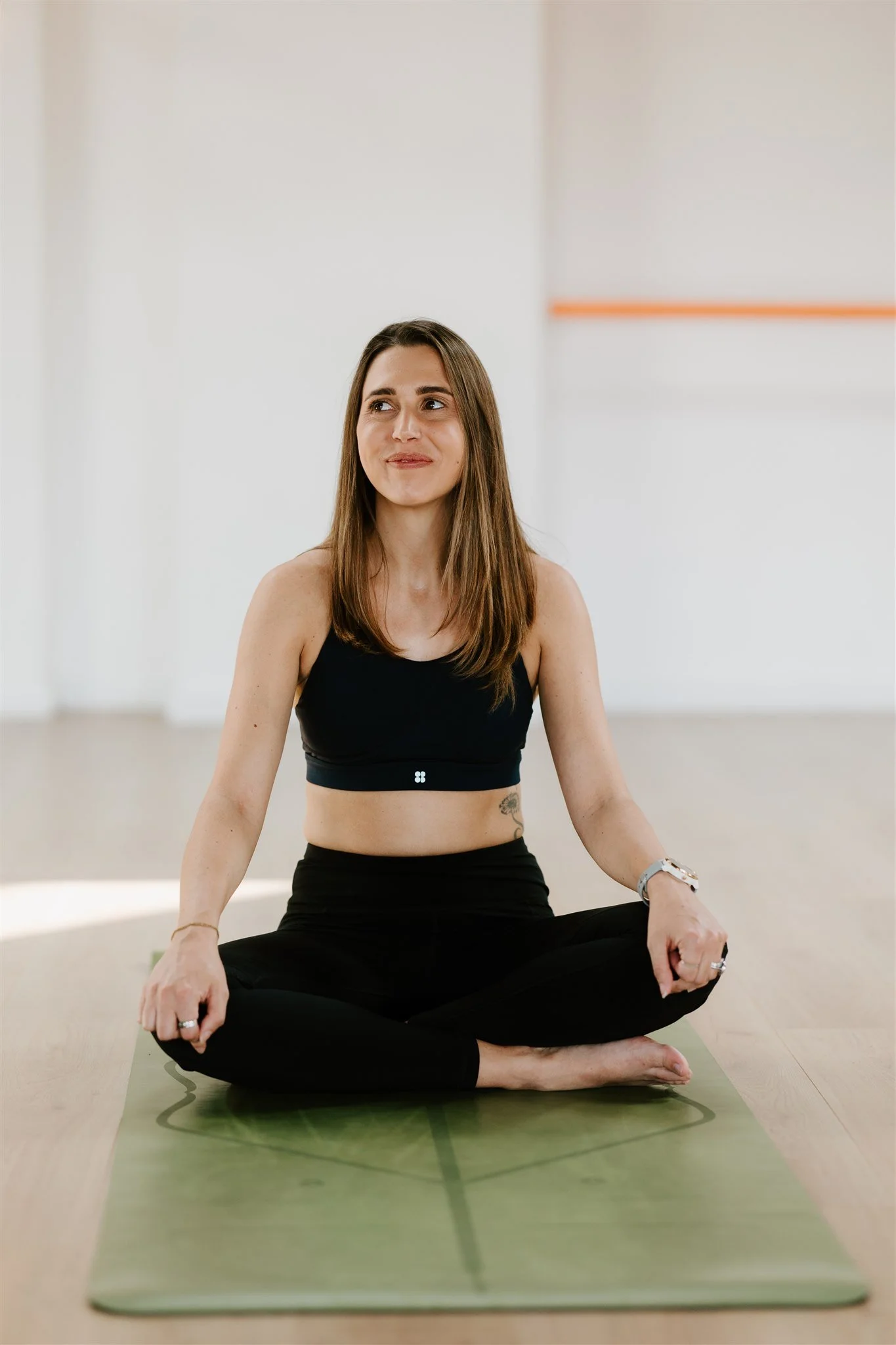 A woman practicing yoga indoors on a green mat, seated cross-legged with hands resting on knees, smiling slightly, wearing a black sports bra and black pants.