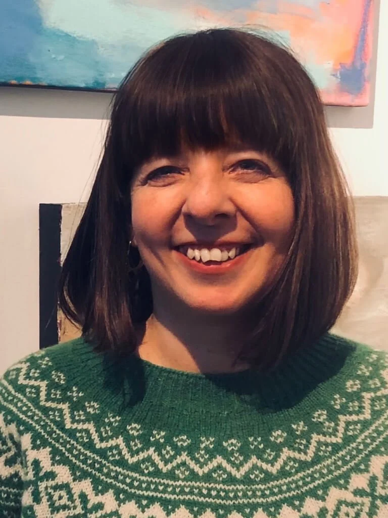 A photo of Maria, smiling with shoulder-length dark brown hair and a fringe, wearing a green jumper with white Nordic pattern, sitting indoors.