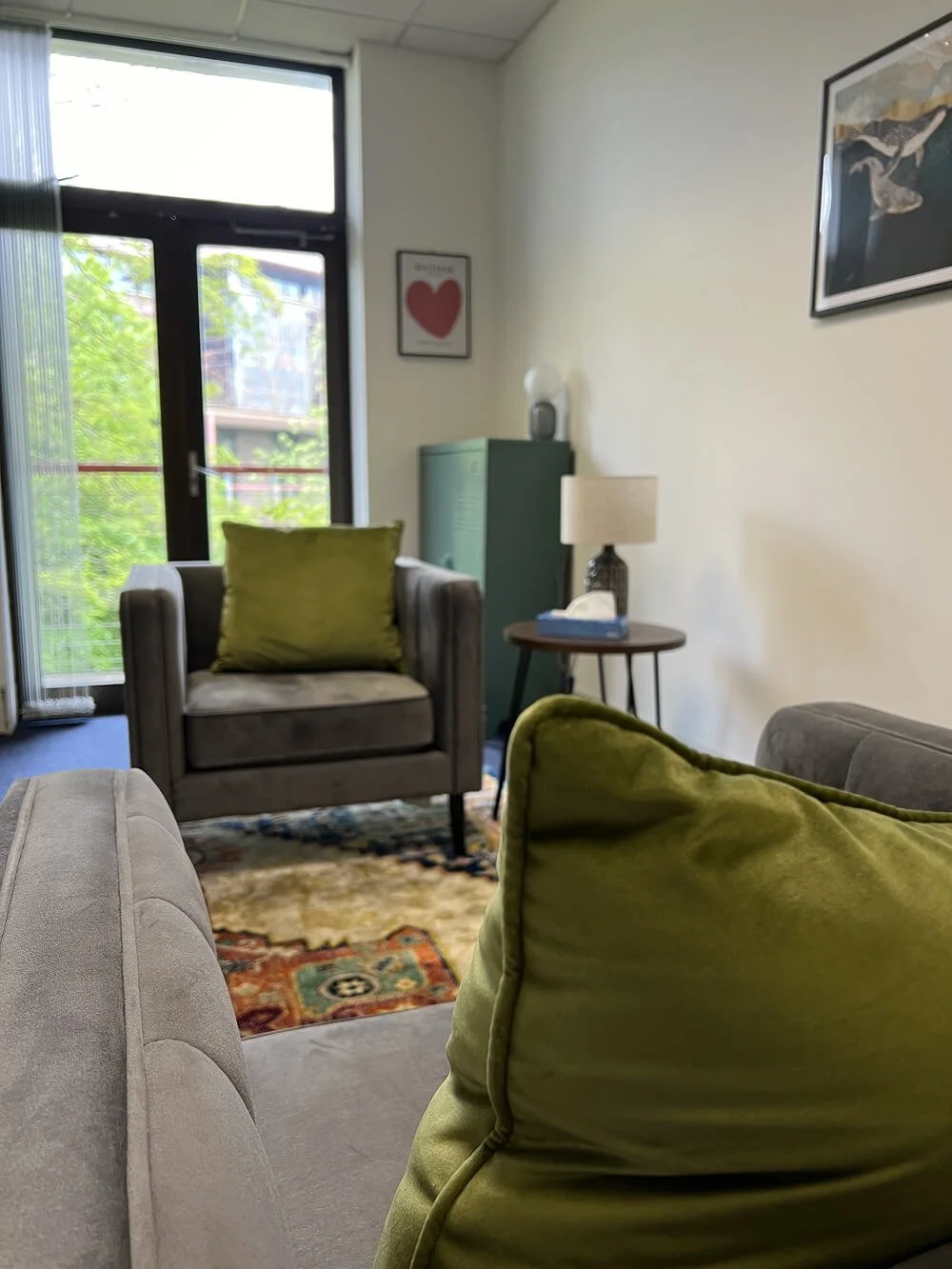 A cozy living room with gray and green accent pillows, a patterned area rug, a gray armchair with a green cushion, a side table with a lamp and tissue box, a green cabinet, framed artwork on the walls, and a glass door leading to a balcony with greenery visible outside.