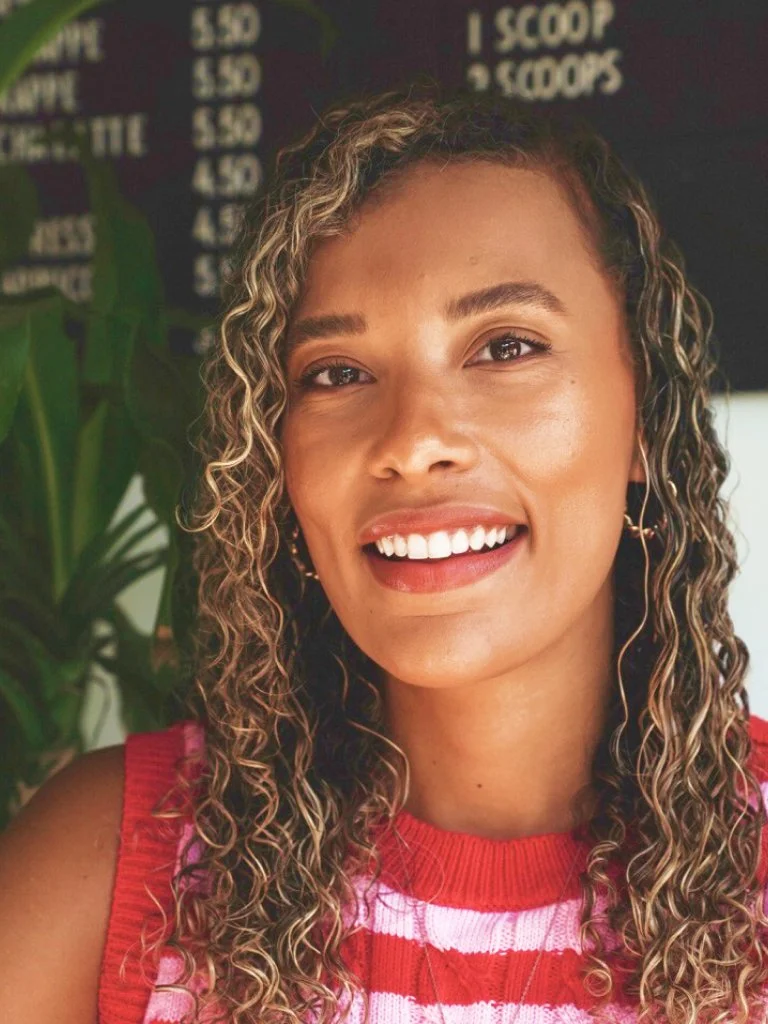 A young woman with curly hair and a bright smile, wearing a red and pink striped sleeveless top, standing indoors near a green plant with a blackboard menu in the background.